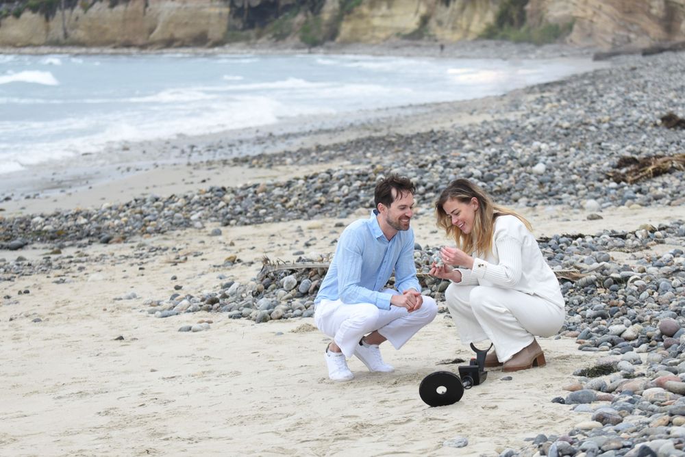 A couple kneels on the rocky beach examining something together near coastal cliffs.