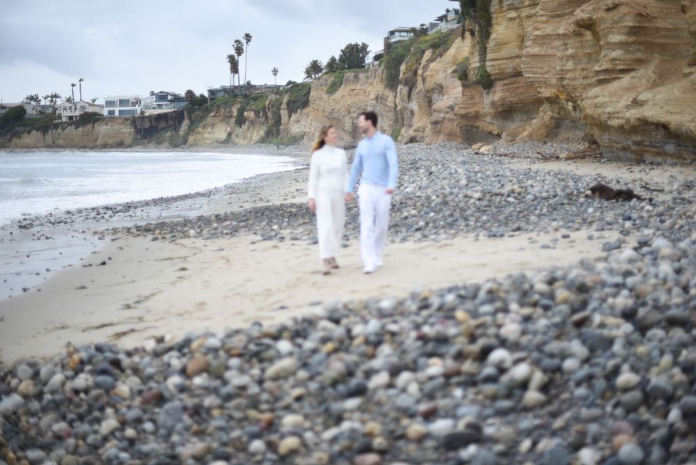 A couple walking hand in hand along a rocky beach with dramatic coastal cliffs behind them.