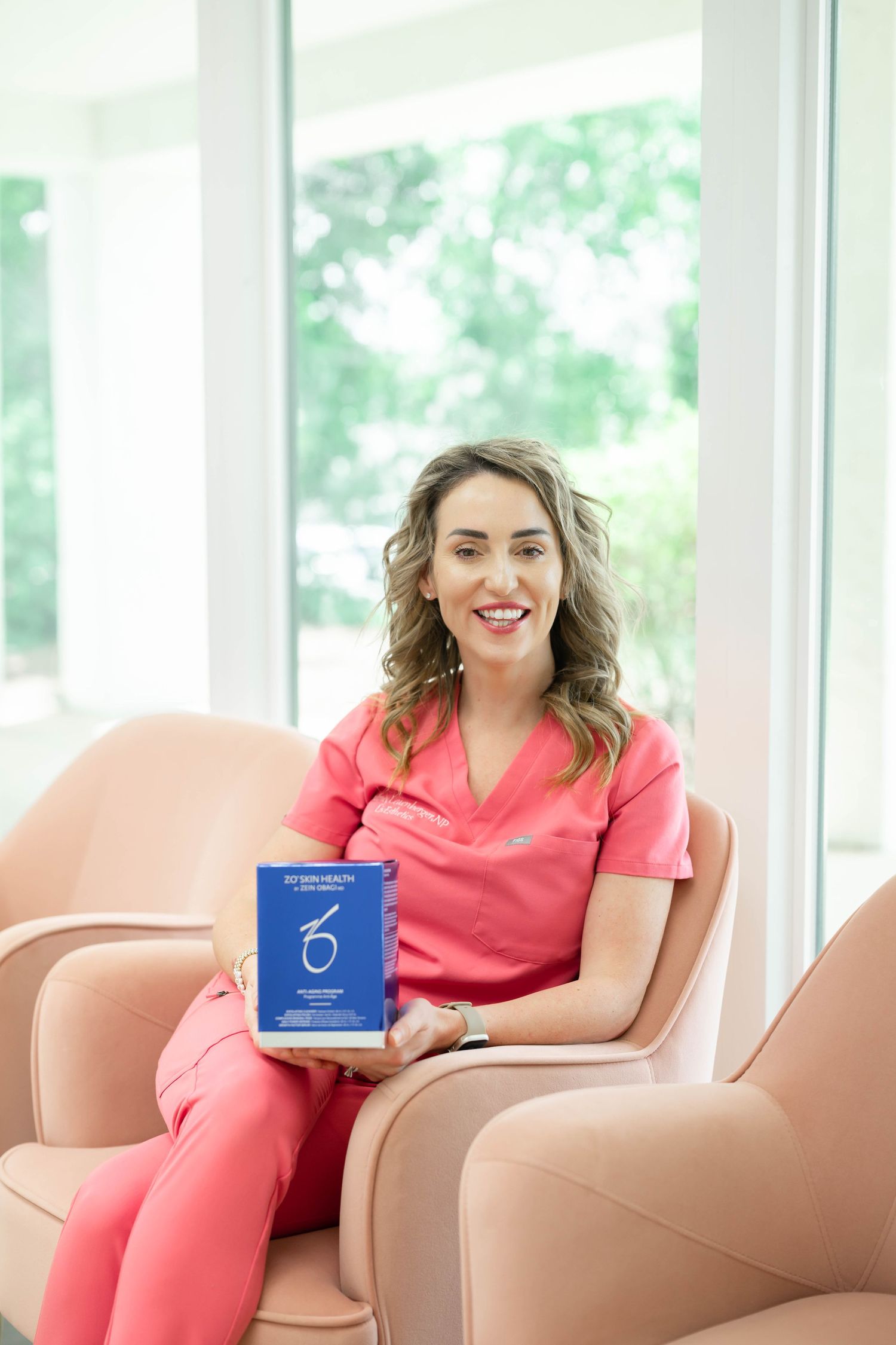 Healthcare professional in coral scrubs sitting in beige chair holding blue medical handbook.