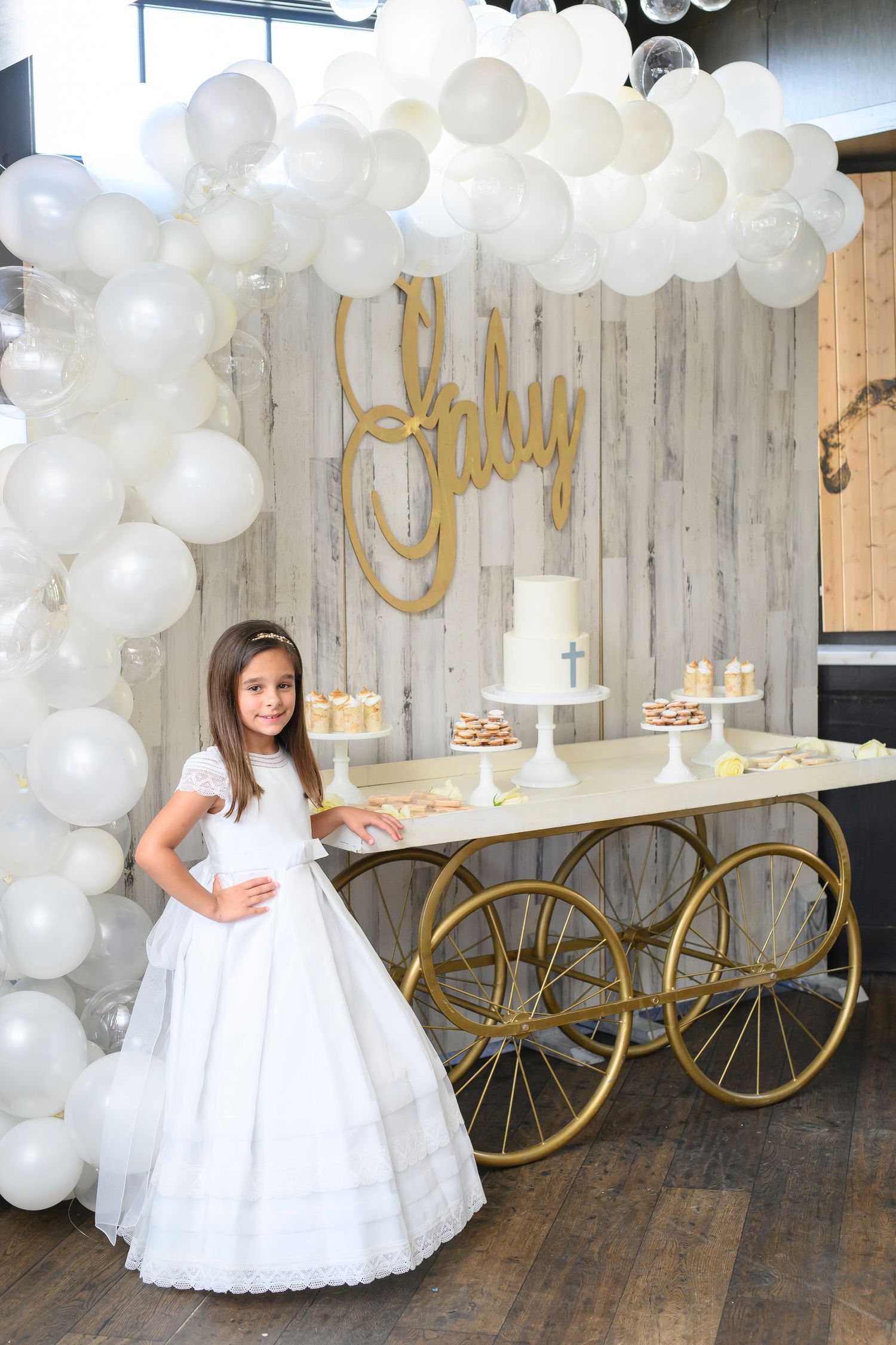 Elegant first communion setup with white balloon arch, gold decorative cart, and dessert display against rustic wood backdrop.