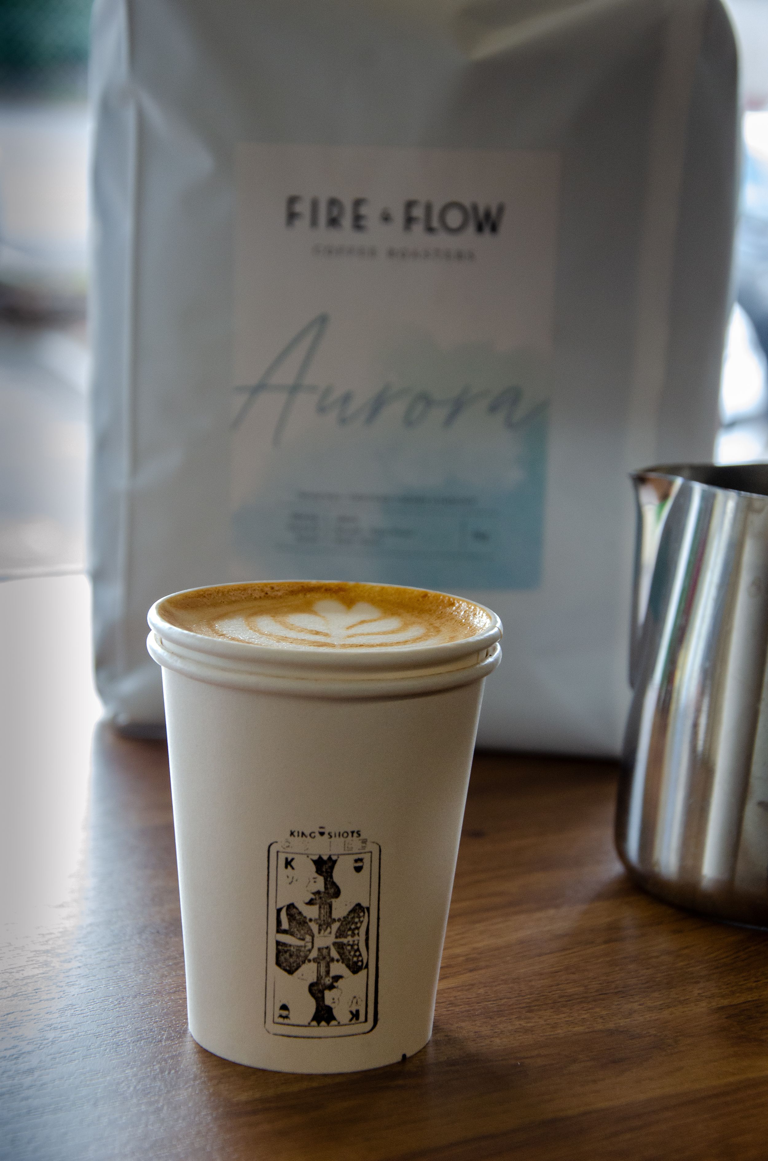 A coffee cup with latte art sits next to a Fire+Flow Aurora coffee bag on a wooden table.