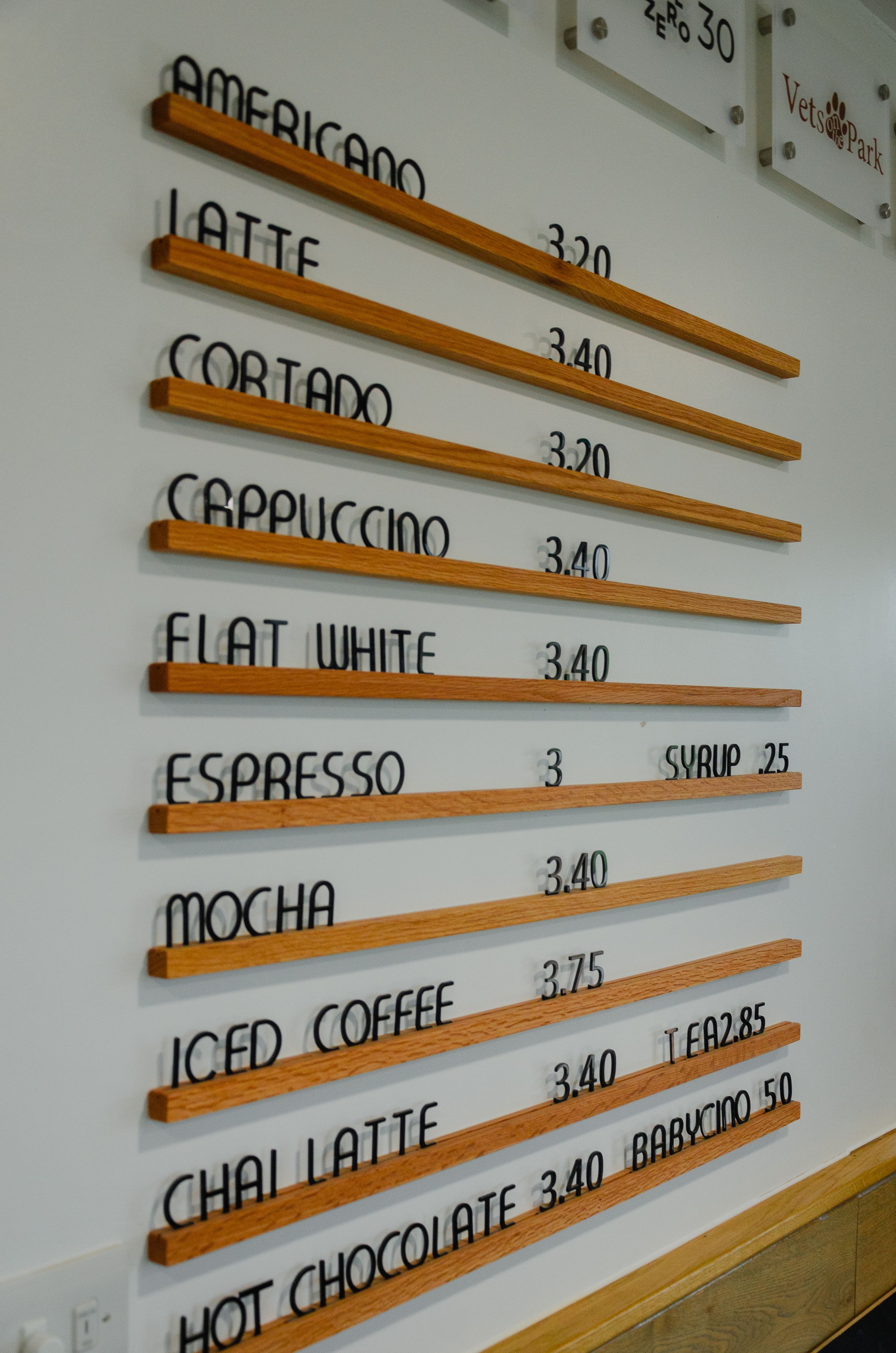 Coffee shop menu board with prices listing various drinks on wooden slats mounted on white wall.