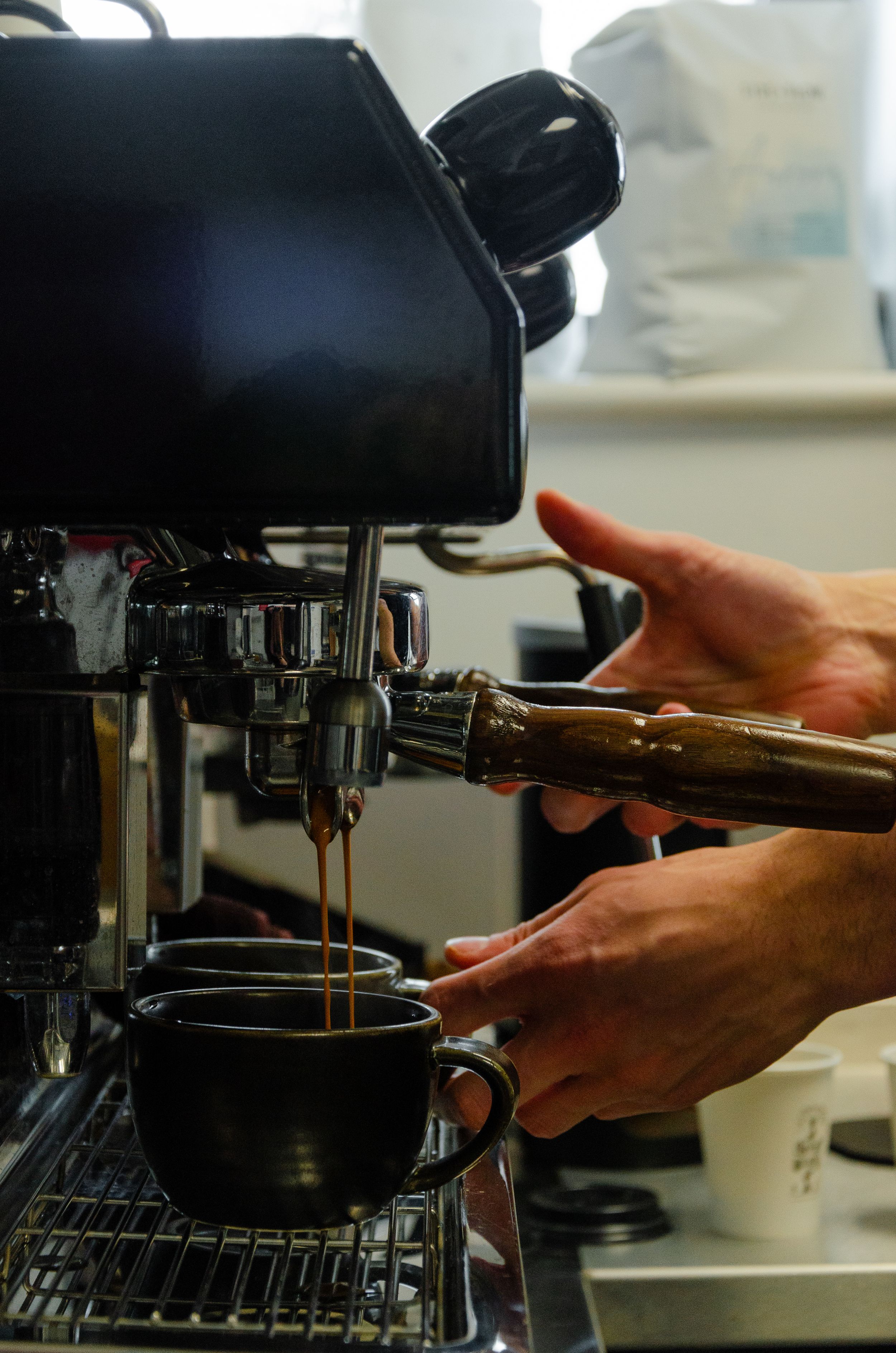 Hands operating an espresso machine while coffee pours into a dark cup.