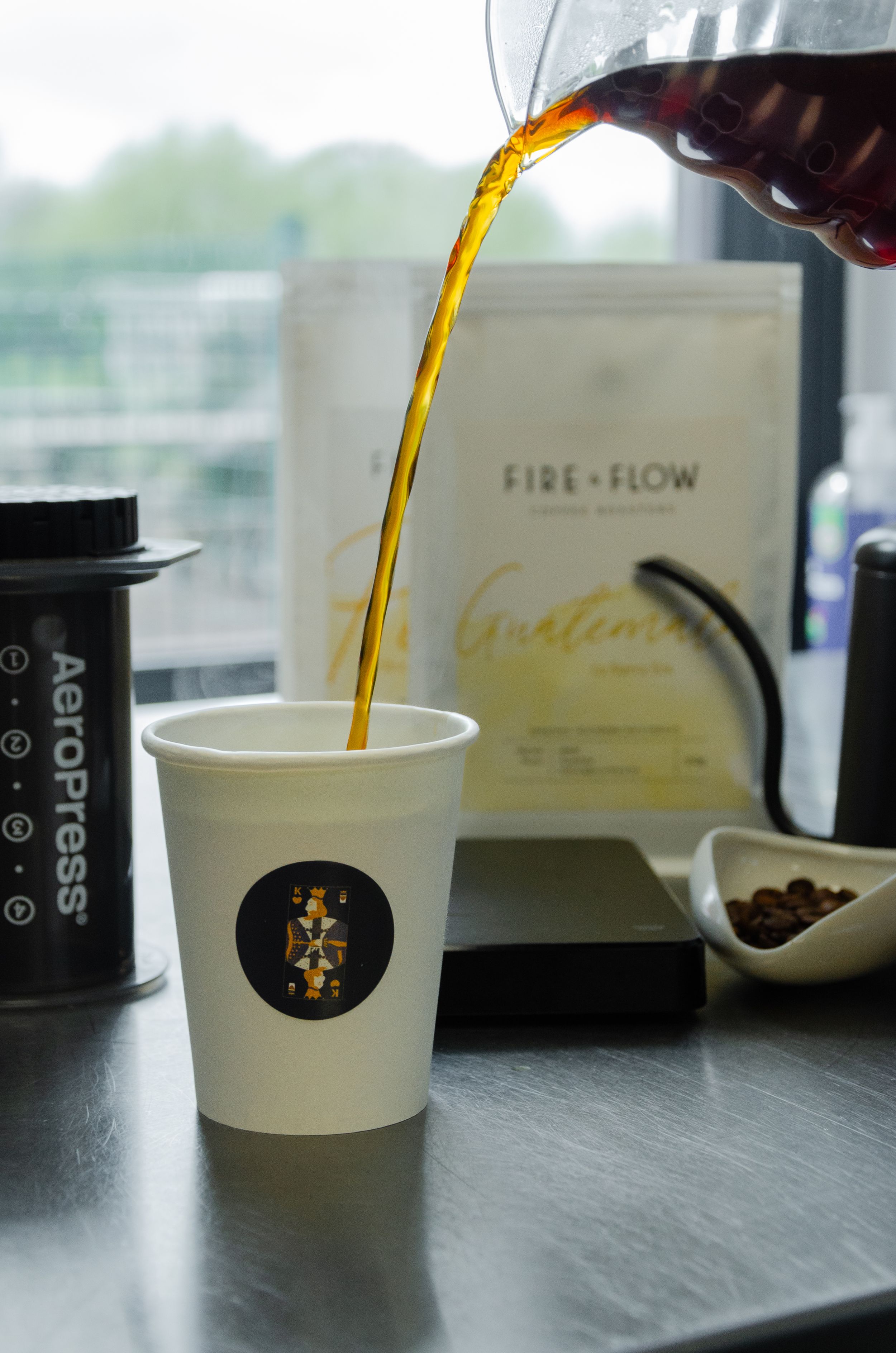 Coffee being poured into a white paper cup with a logo against a bright window background.