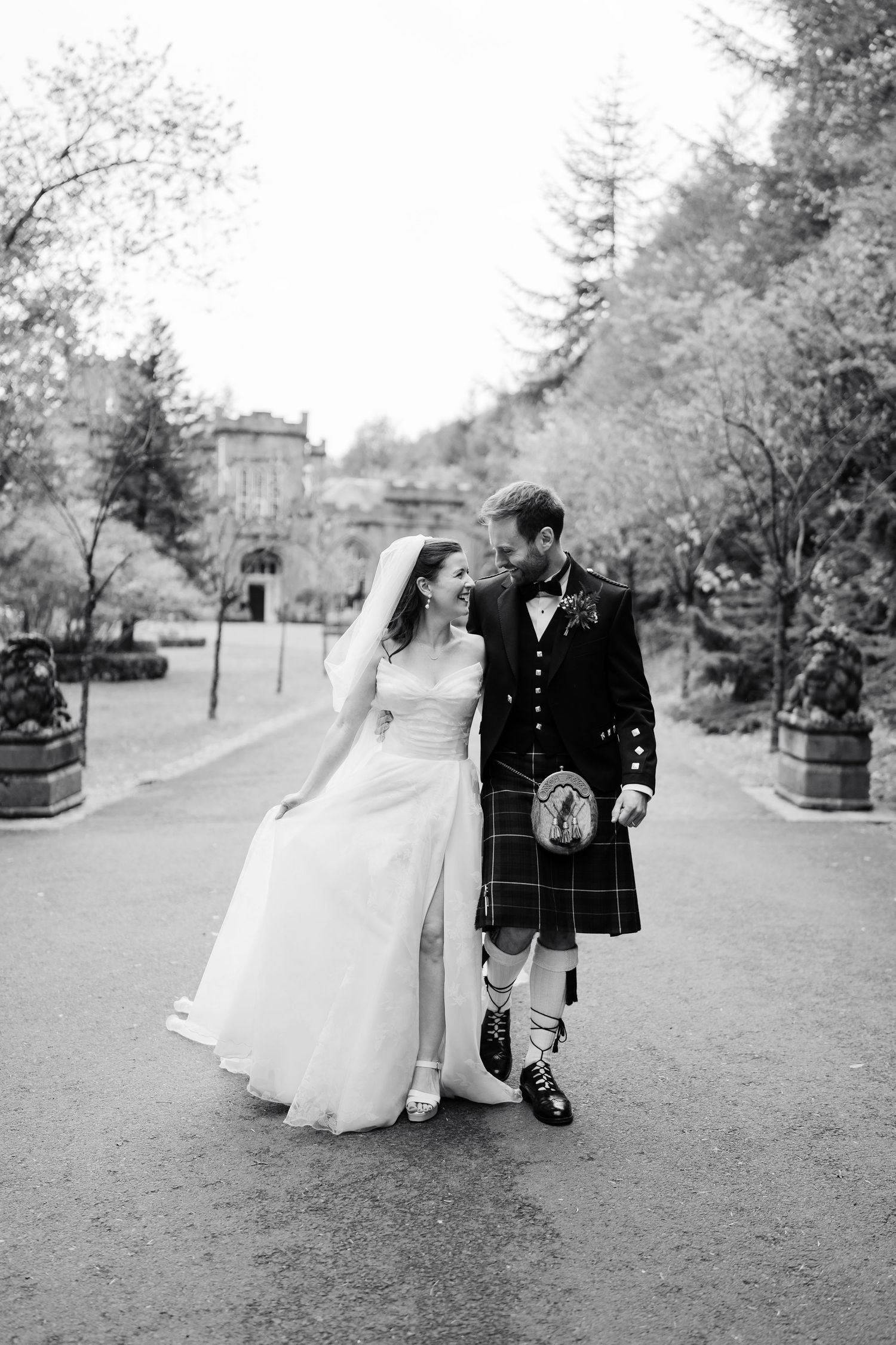 A couple in wedding attire walks along a tree-lined path at a Scottish estate.