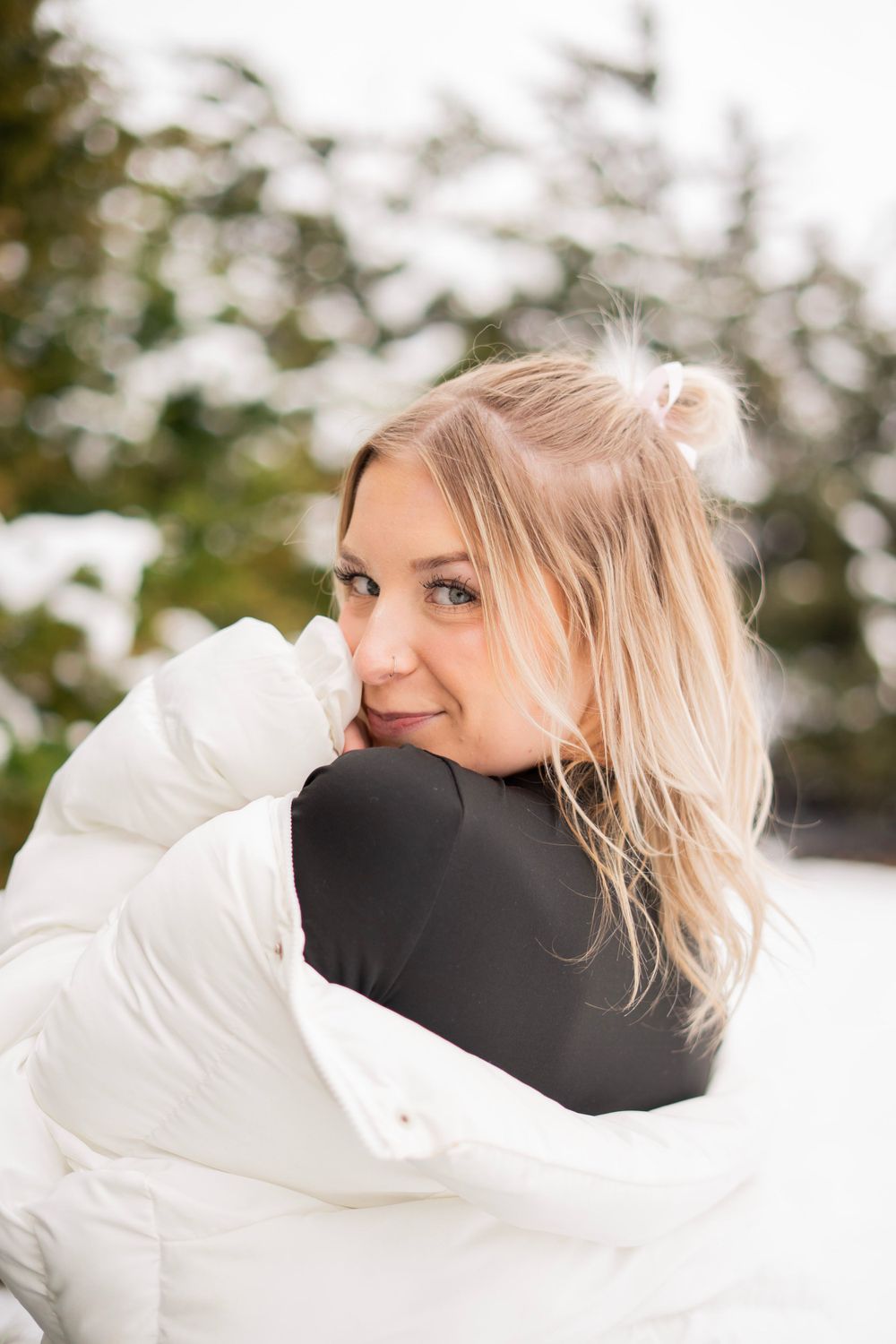 Smiling blonde woman in white puffer jacket posing outdoors in snowy winter setting.