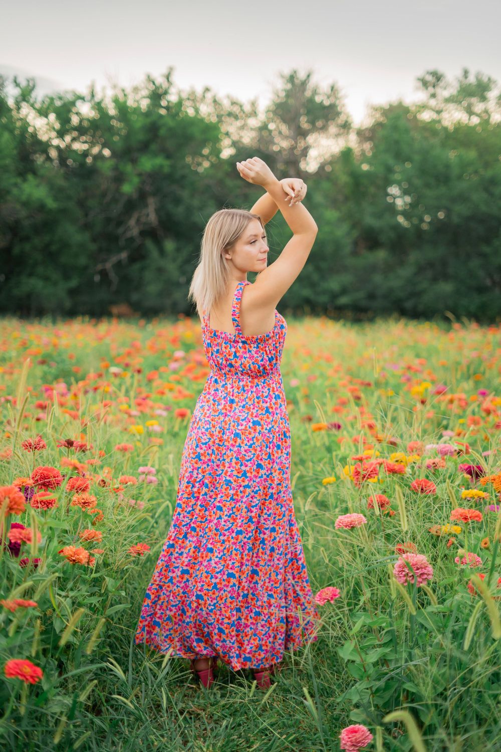 Blonde girl in floral maxi dress standing with arms raised in a colorful zinnia field.