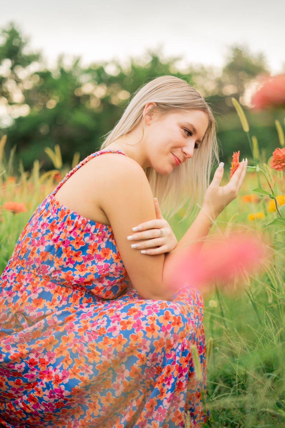 Blonde teen girl in floral dress smiling while touching flowers in a colorful garden.