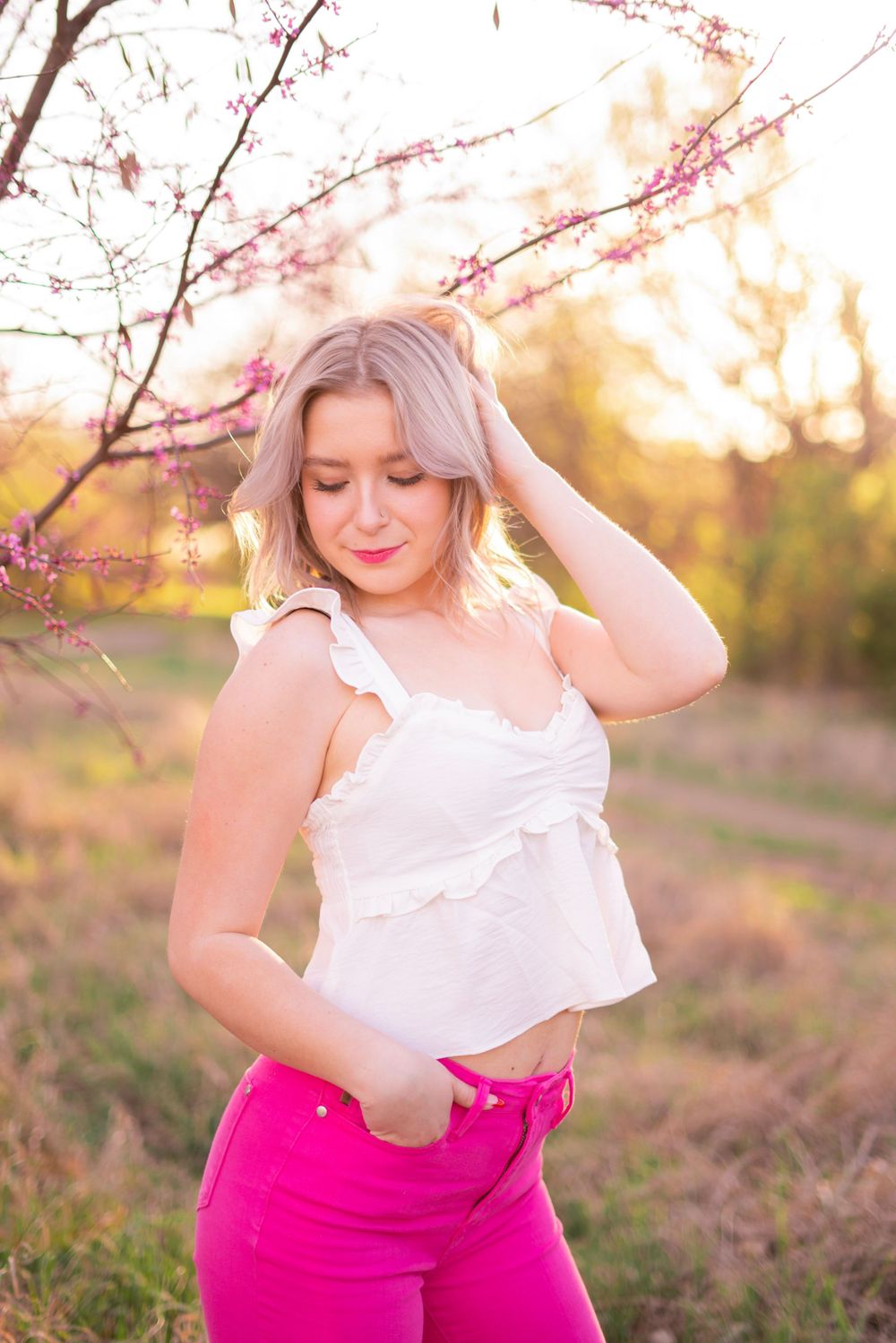 Blonde young woman in white top and pink jeans posing near pink blossoming trees at sunset.