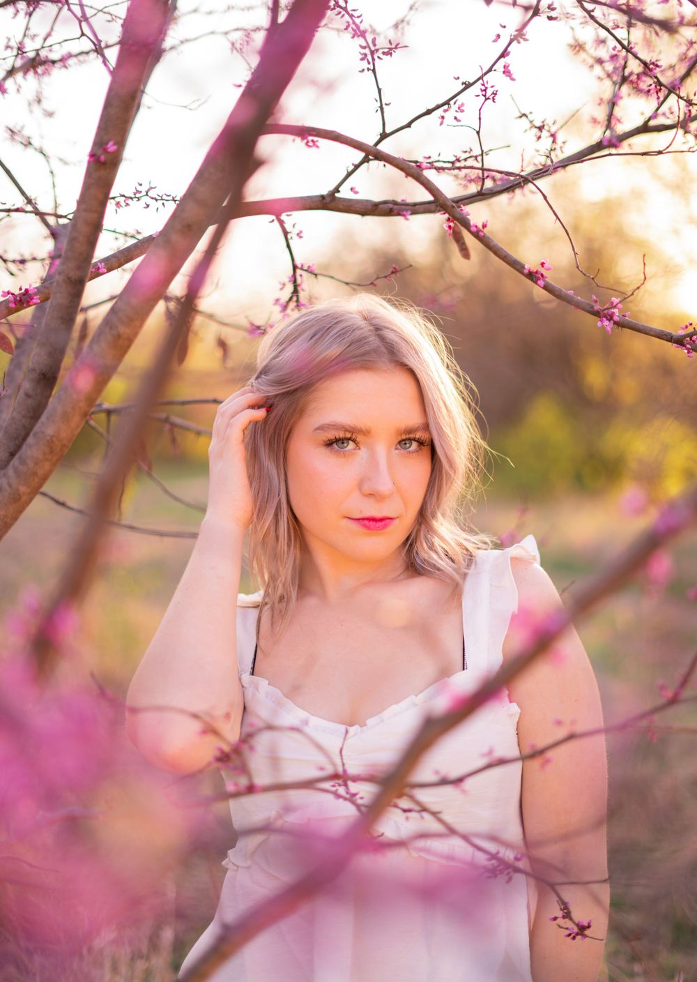 Young blonde woman in white dress surrounded by pink spring blossoms at golden hour.