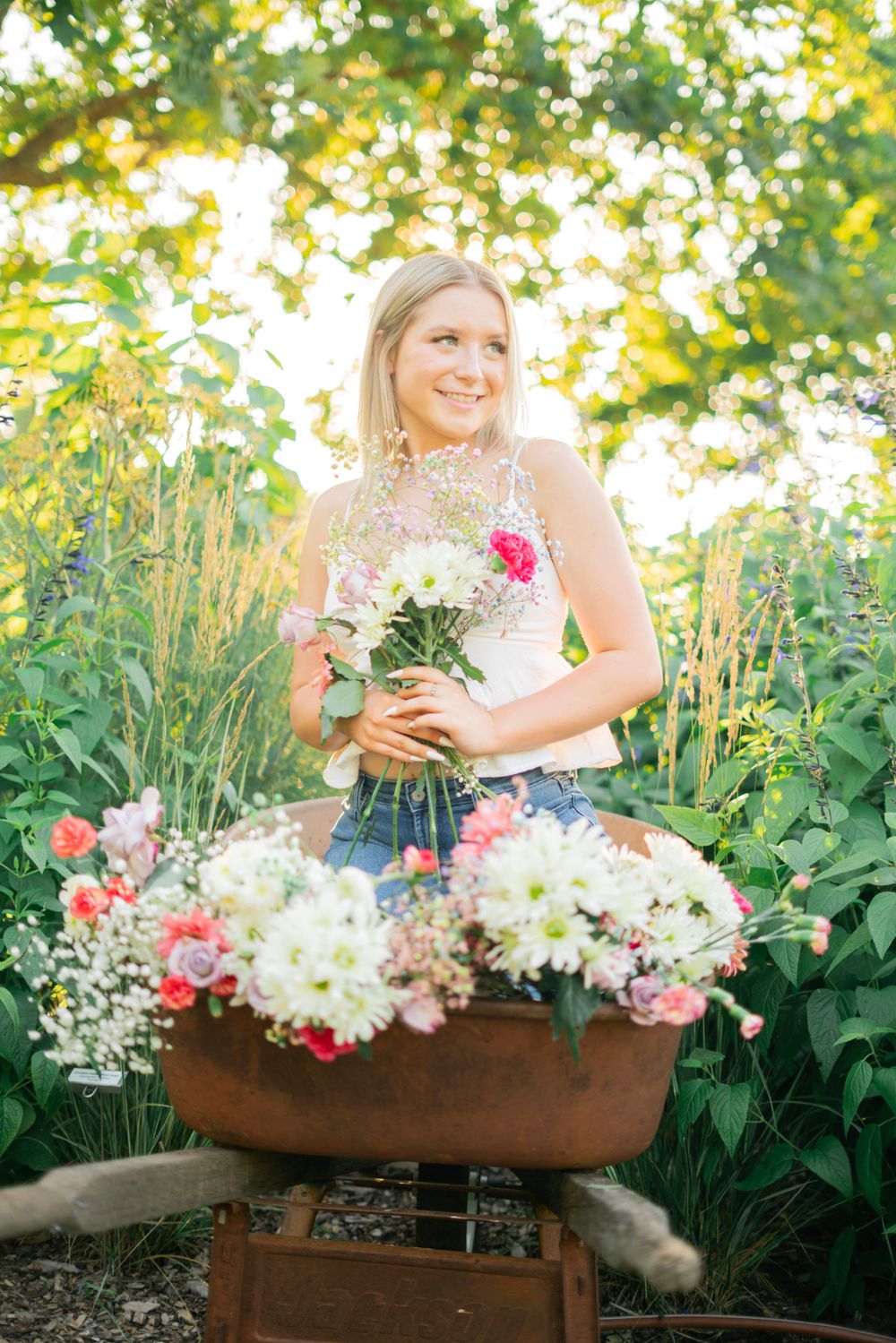 Smiling blonde teen girl holding flowers in a rustic tub surrounded by lush garden greenery.