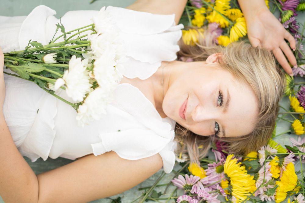 Blonde woman lying among vibrant yellow and purple wildflowers holding white blooms softly.
