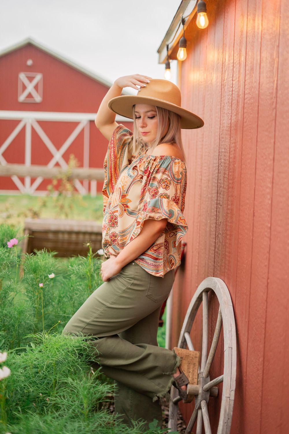 Blonde girl in boho floral top and hat leaning against red barn with wagon wheel.