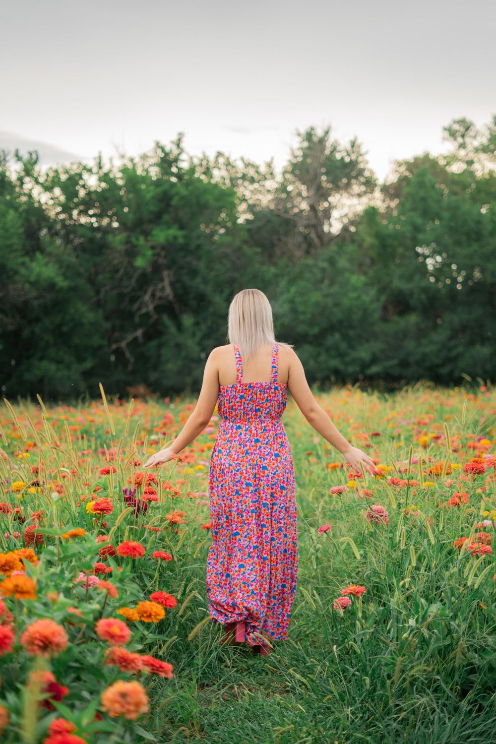 Blonde woman in floral maxi dress walking through vibrant field of orange and red zinnia flowers.