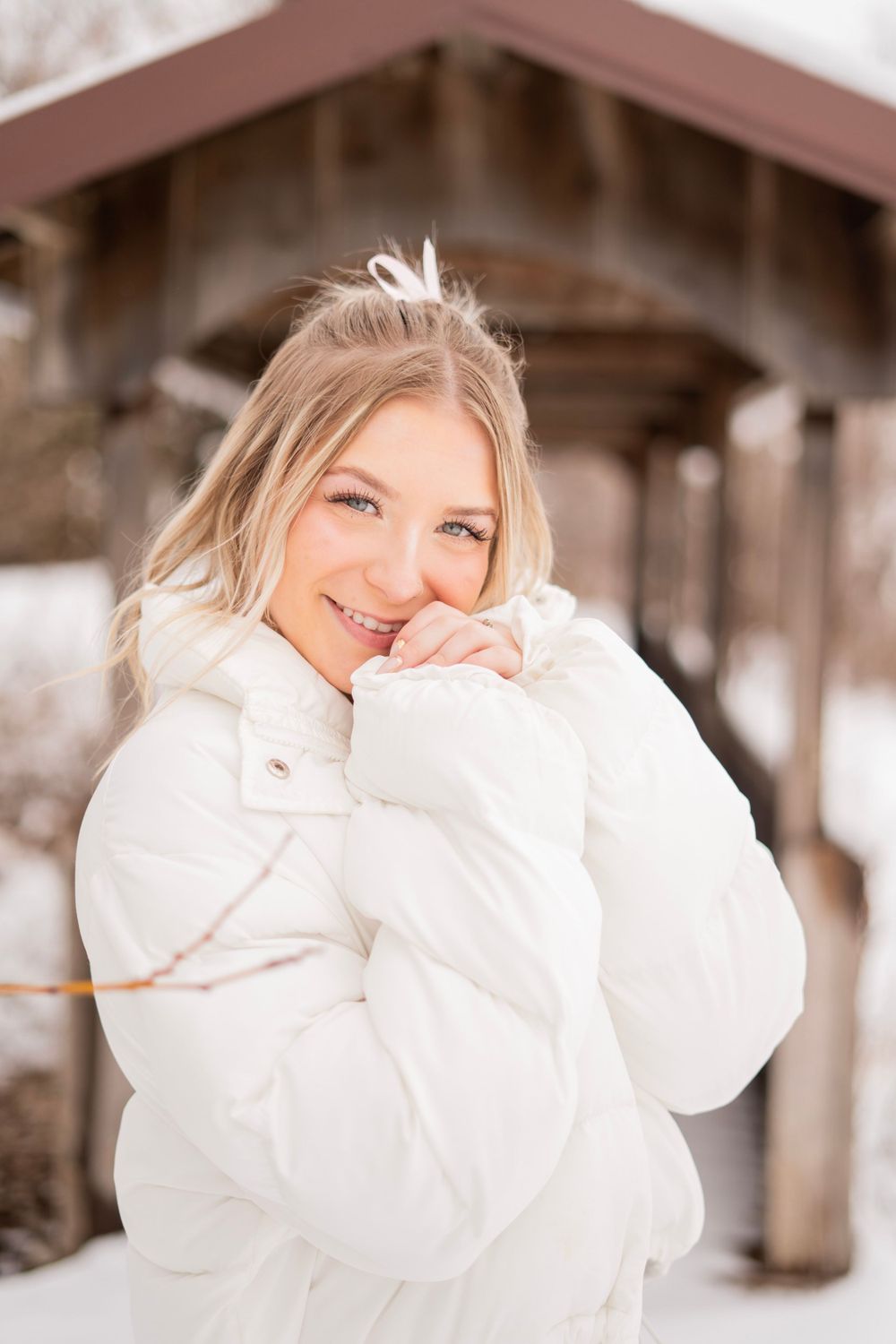 Cheerful blonde woman in white puffer coat posing near wooden structure in snowy landscape.