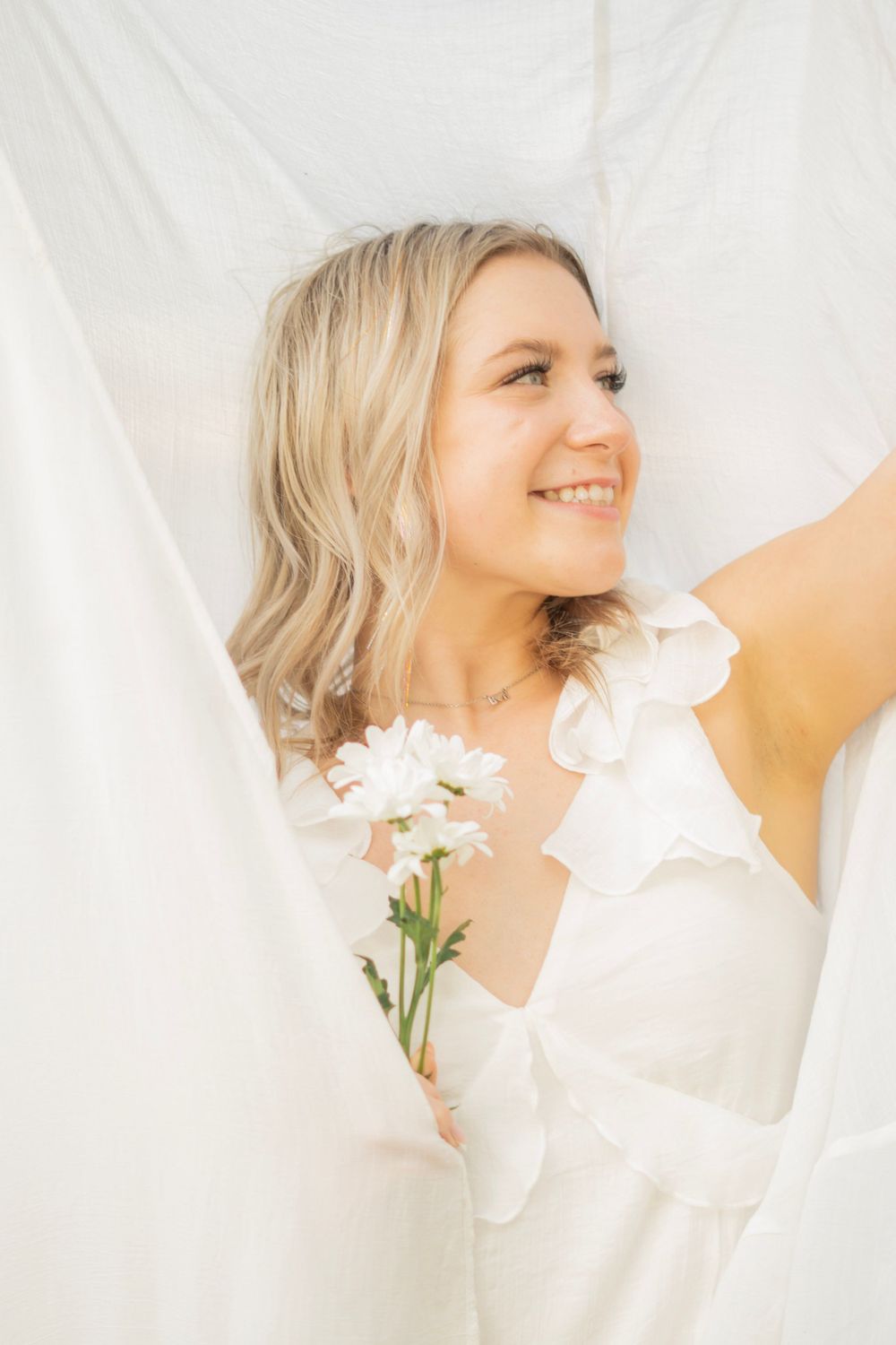 Blonde woman in white ruffled dress holding white flowers against a soft white backdrop.