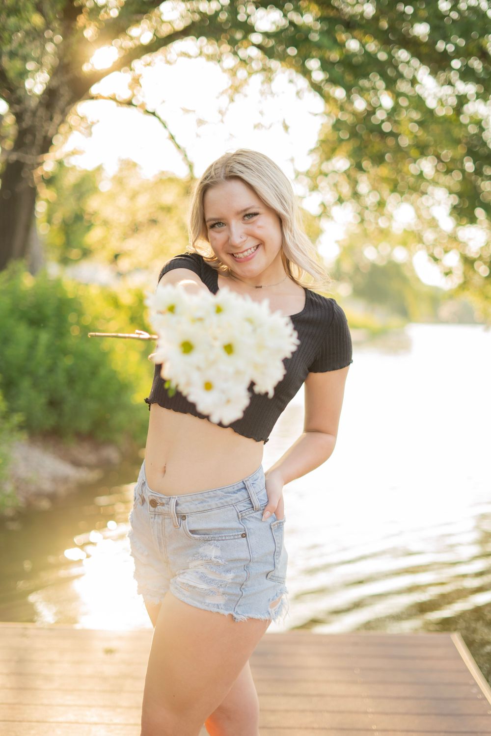 Smiling blonde woman holding white daisy bouquet on a sunny lakeside wooden dock.