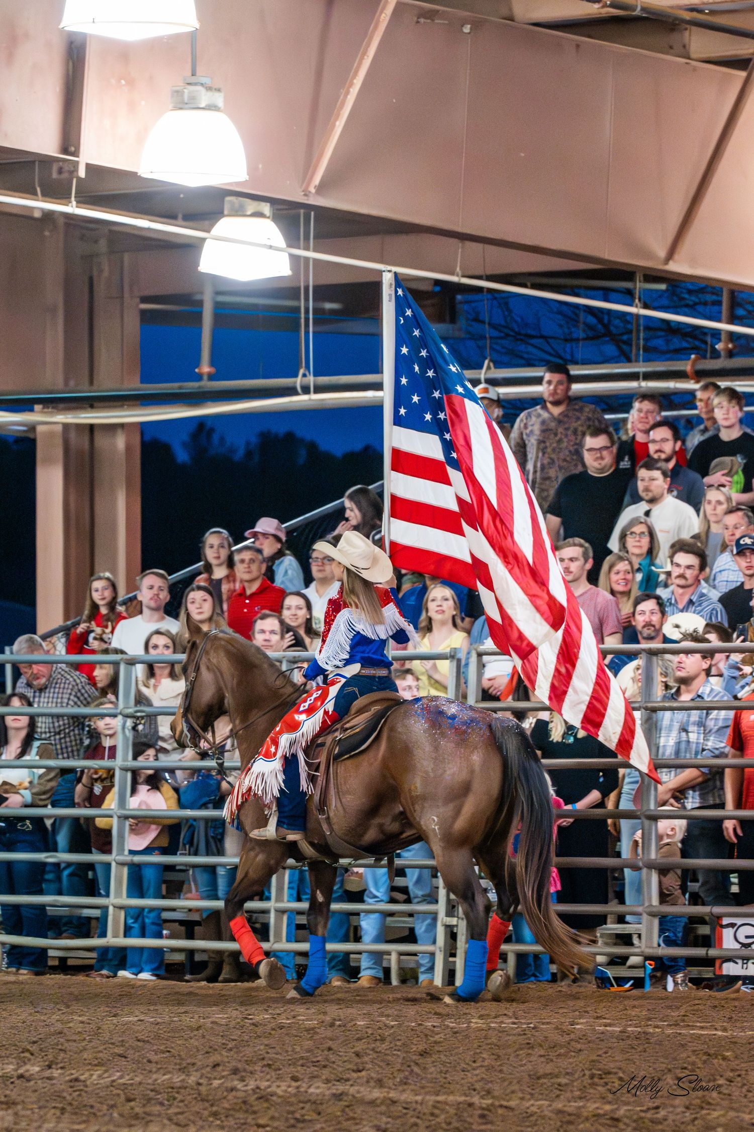 Rodeo rider on horseback carrying a large American flag before a packed crowd at an indoor arena.