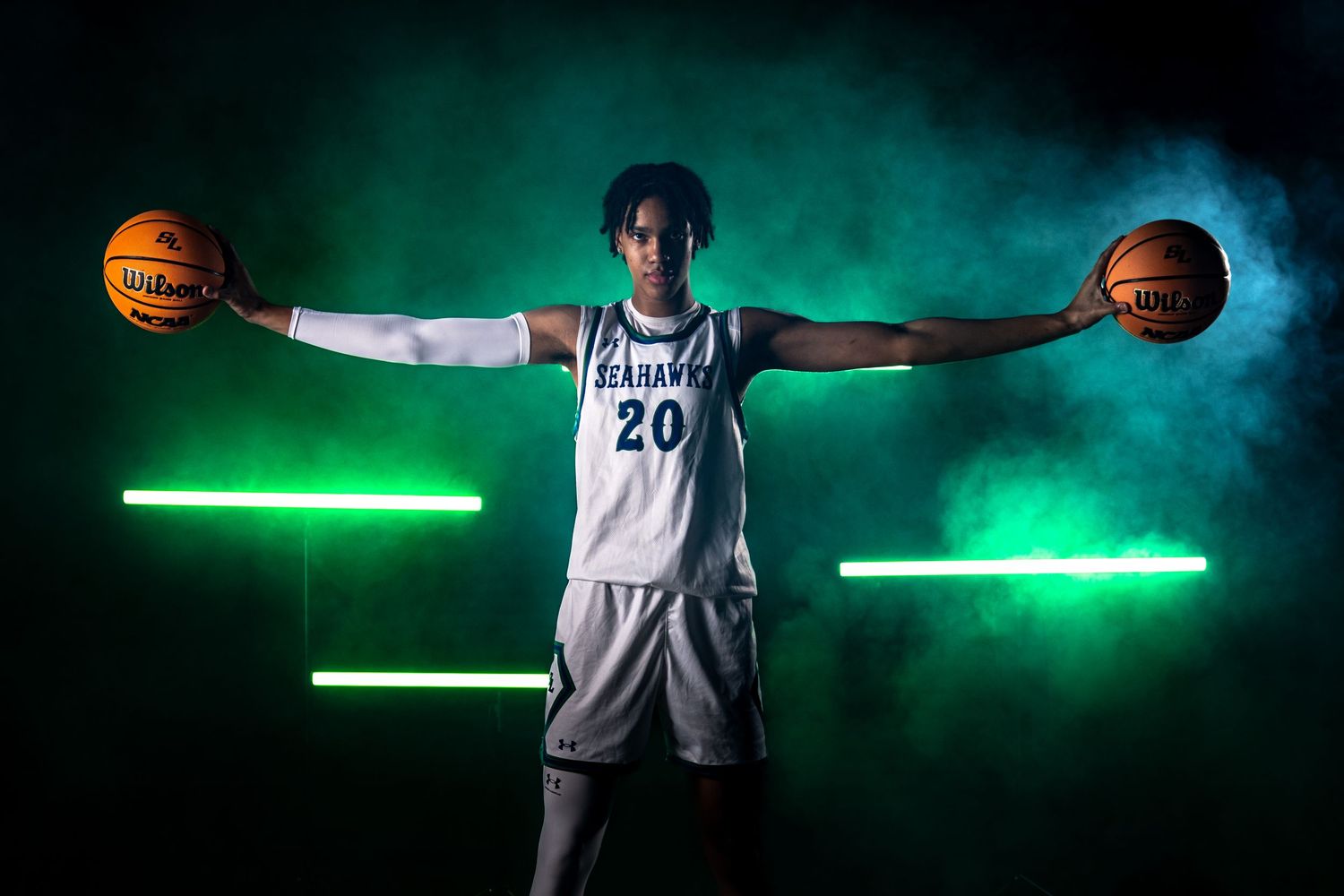 Basketball player holds two basketballs outstretched in dramatic pose with green lighting effects in the background. sports Media day
