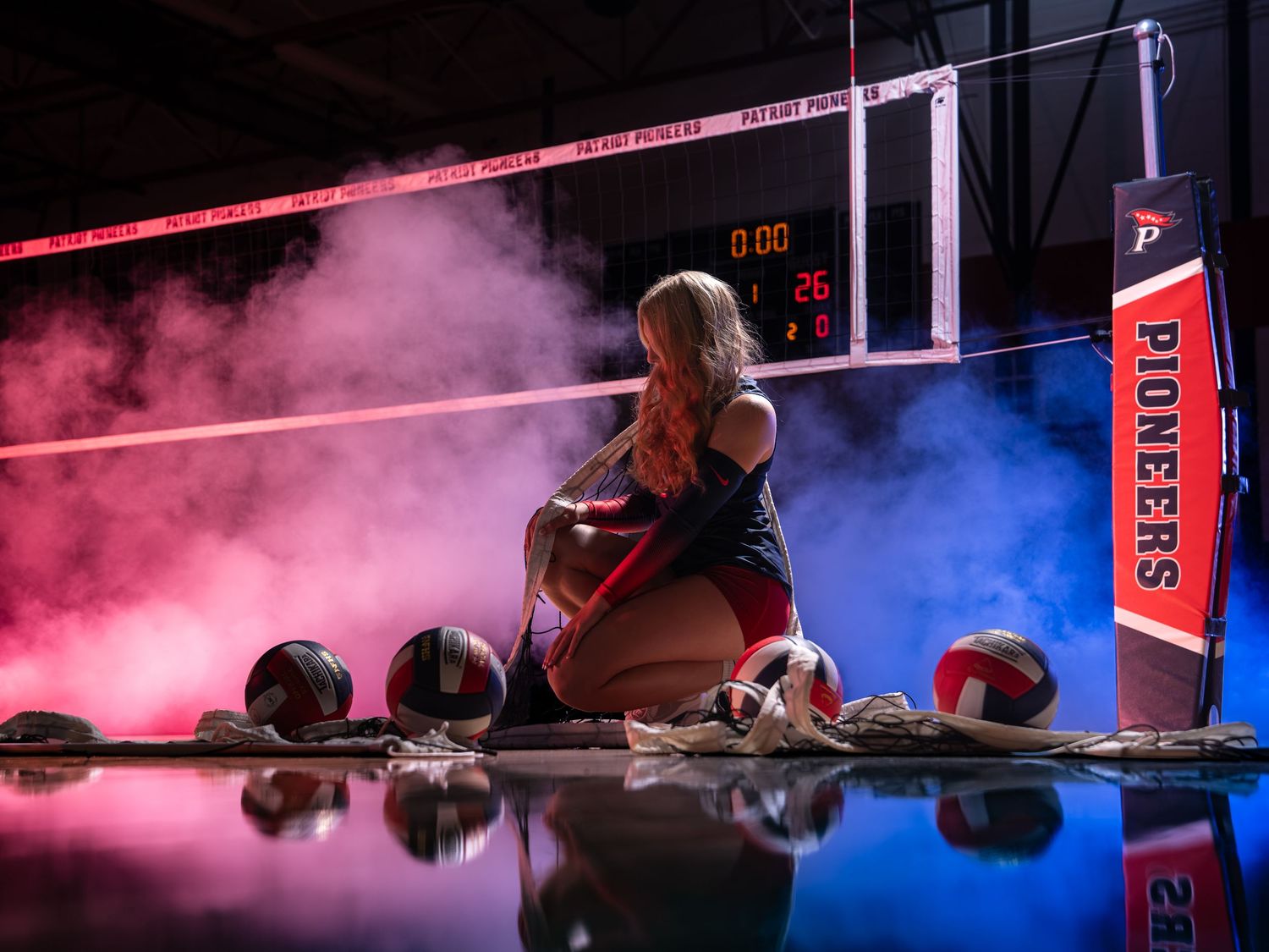 Female volleyball player sits surrounded by balls in smoky colorful gymnasium setting.