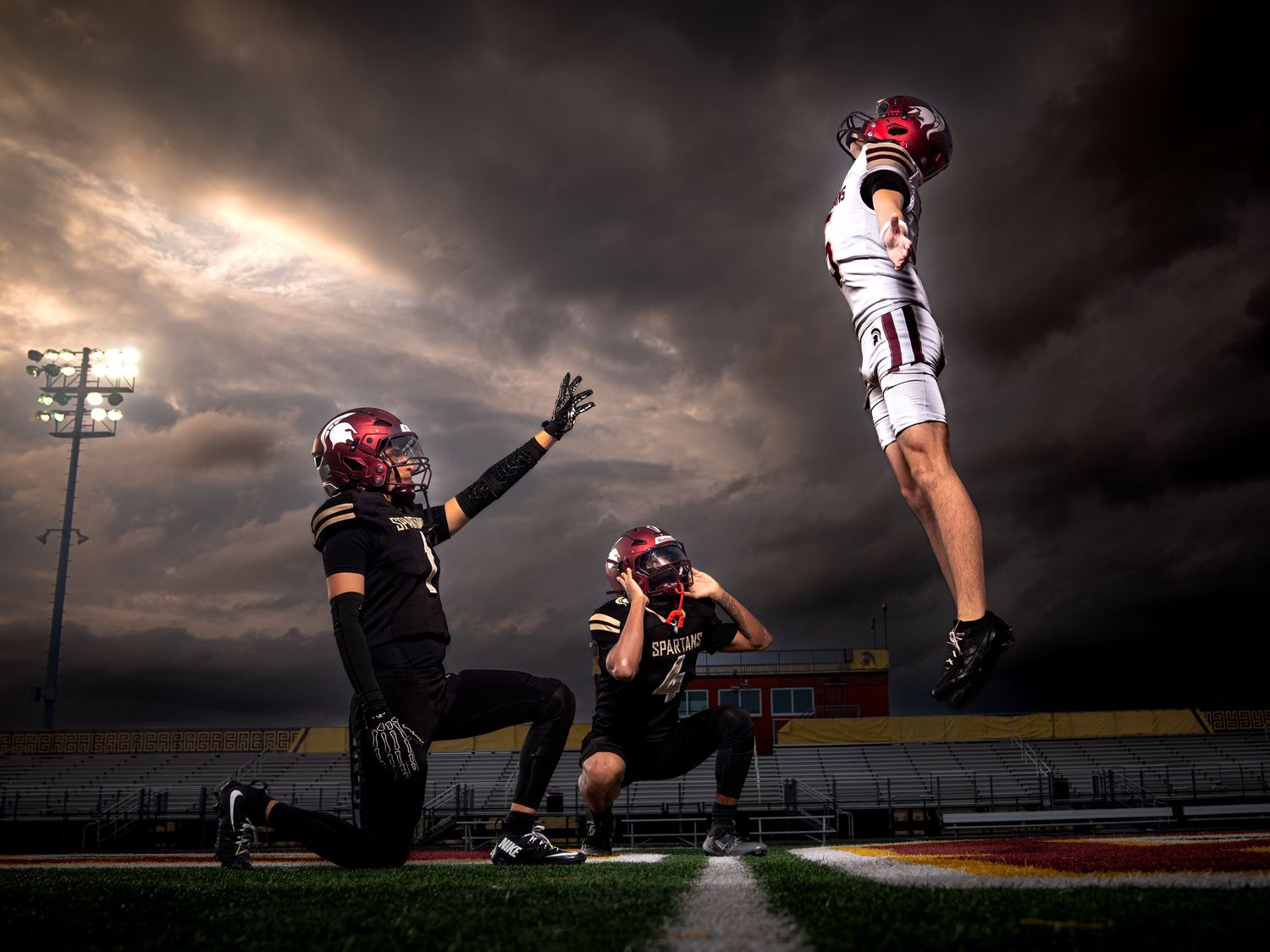 Three Spartans football players perform dynamic jump pose under dramatic storm clouds.