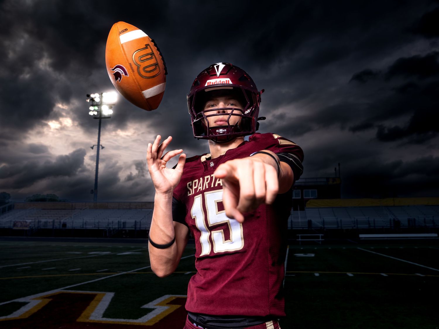 Spartans football player in maroon jersey tosses ball under dramatic stormy sky.