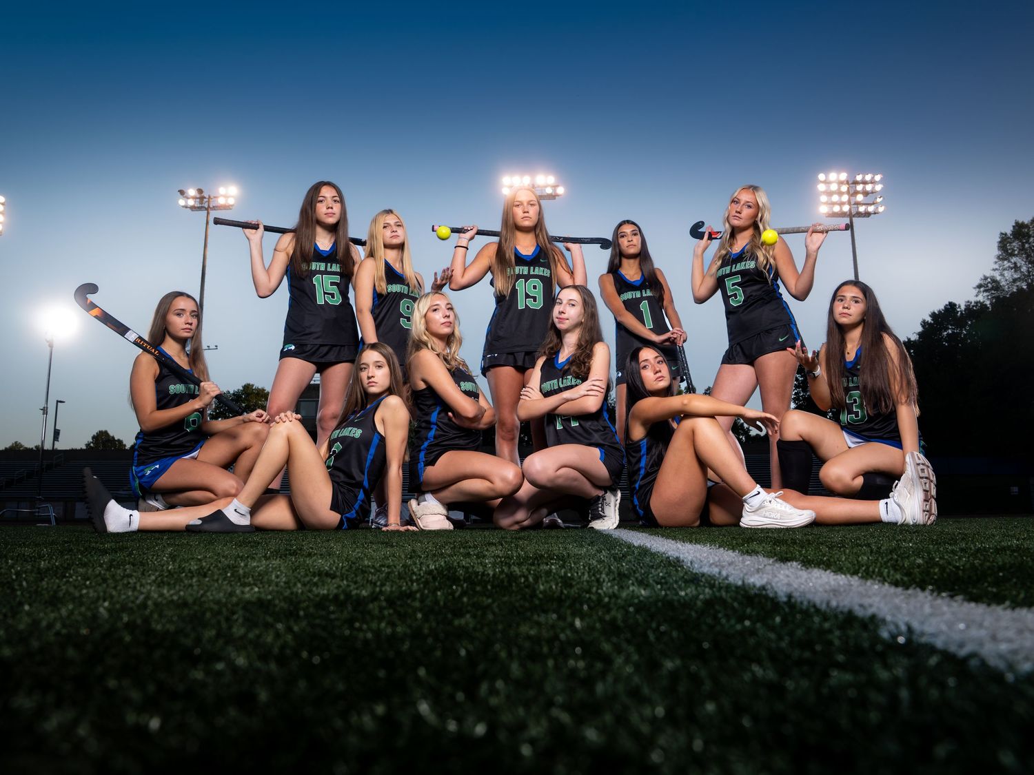 Girls field hockey team poses dramatically under stadium lights at dusk holding sticks.
