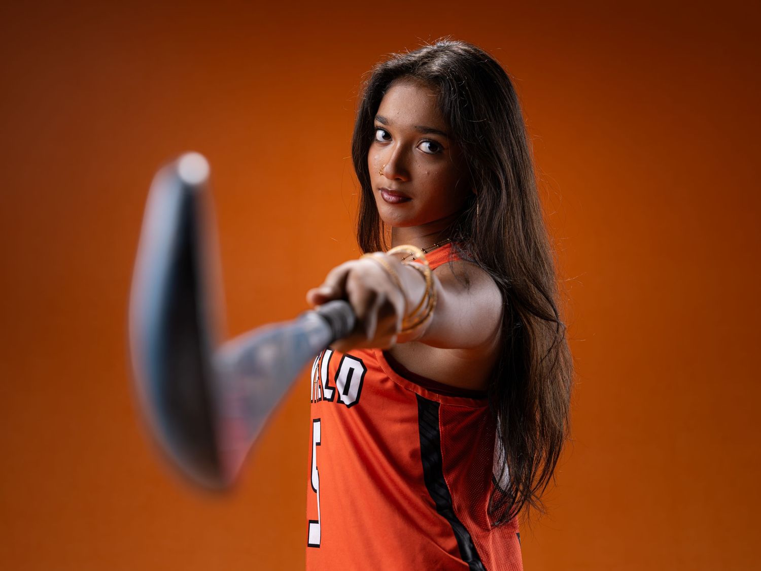 Female athlete in orange jersey extends hockey stick toward camera in dynamic sports portrait.