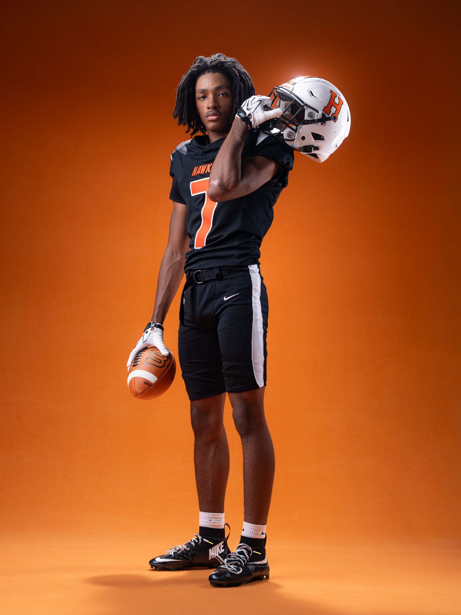 High school football player in black and orange Nike uniform holds helmet and football on orange background.