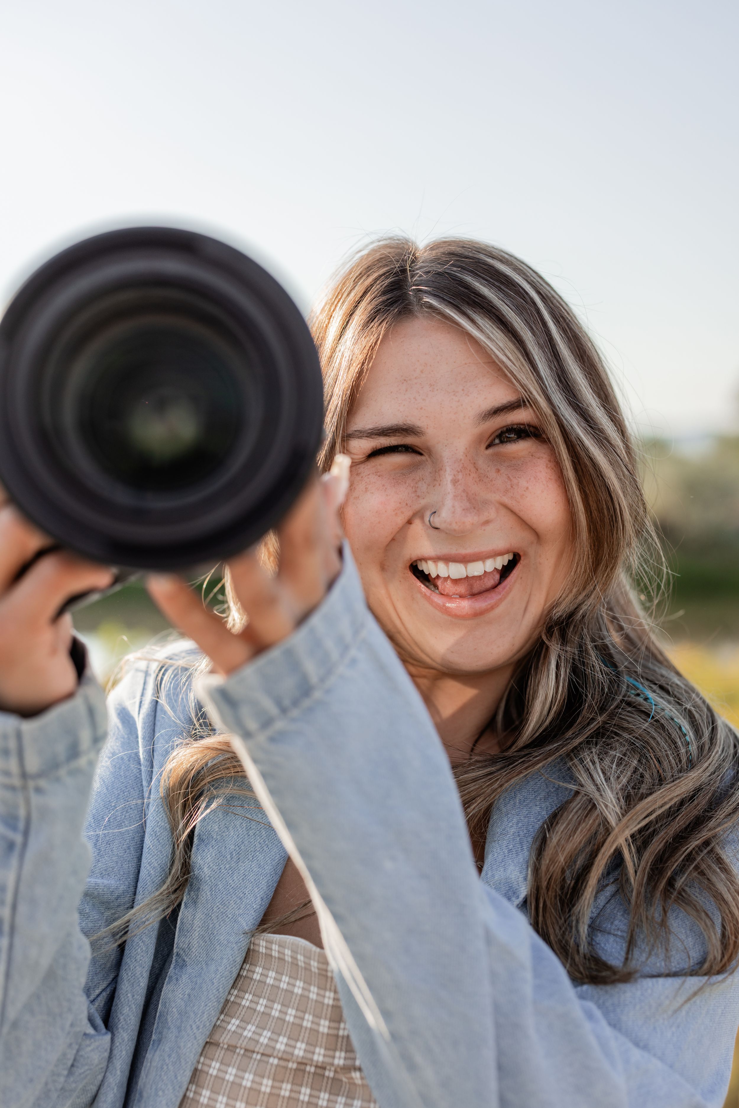 Smiling young woman with long blonde hair laughing outdoors while a photographer captures her portrait.