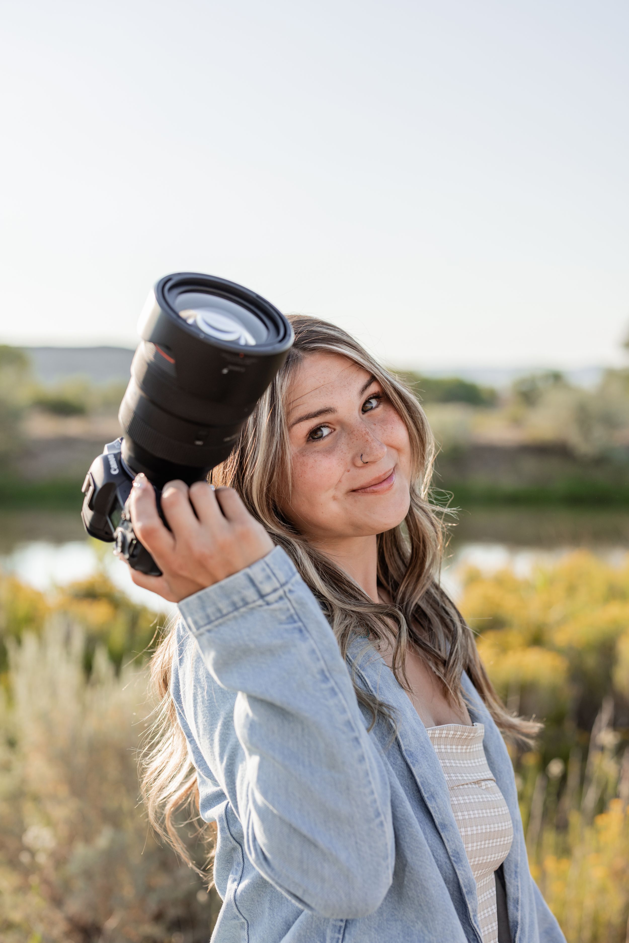 Young woman with blonde hair holding a DSLR camera outdoors, wearing a denim jacket, smiling in natural light.