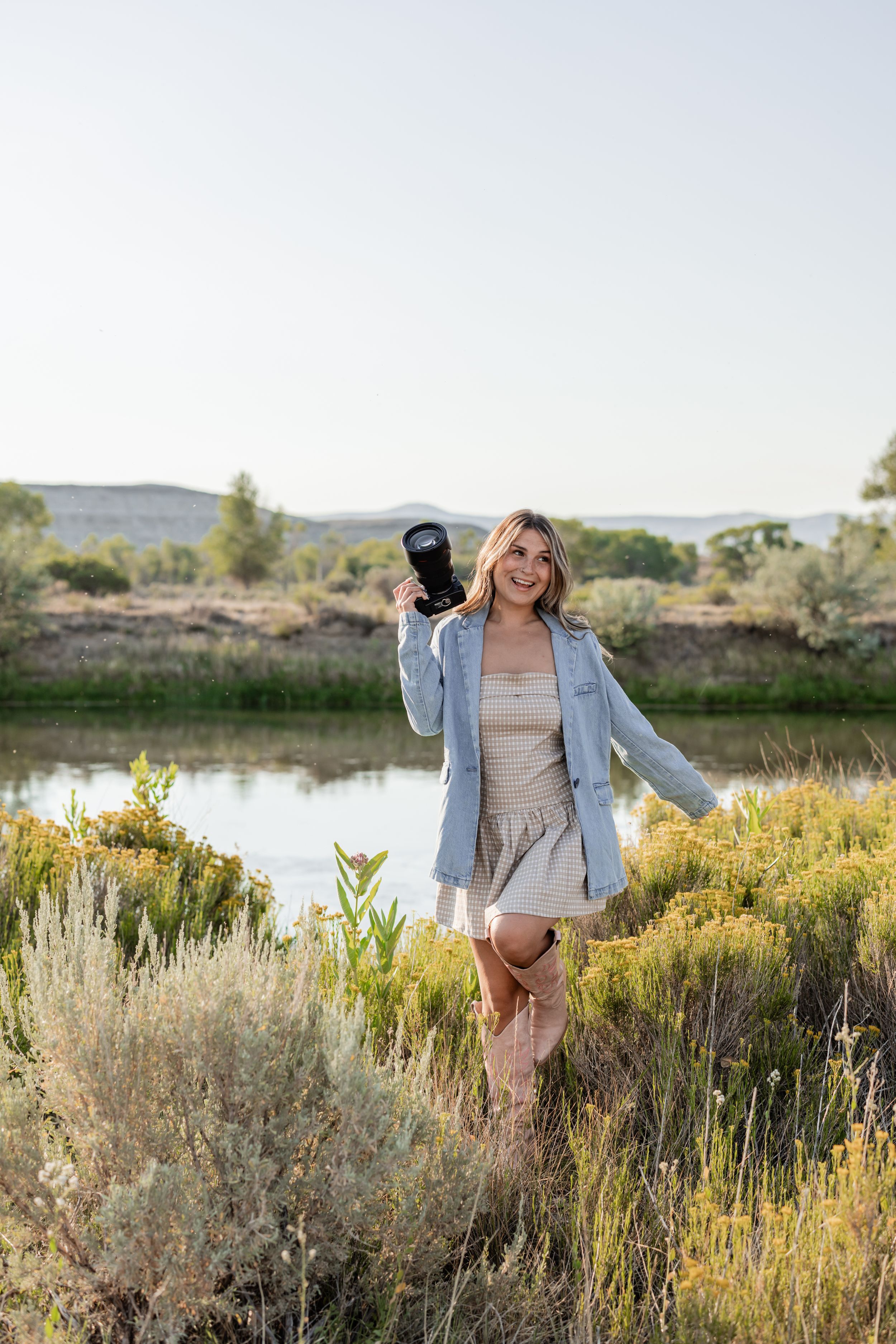 Smiling blonde woman in denim jacket and mini dress holds camera aloft near a scenic river with lush greenery.