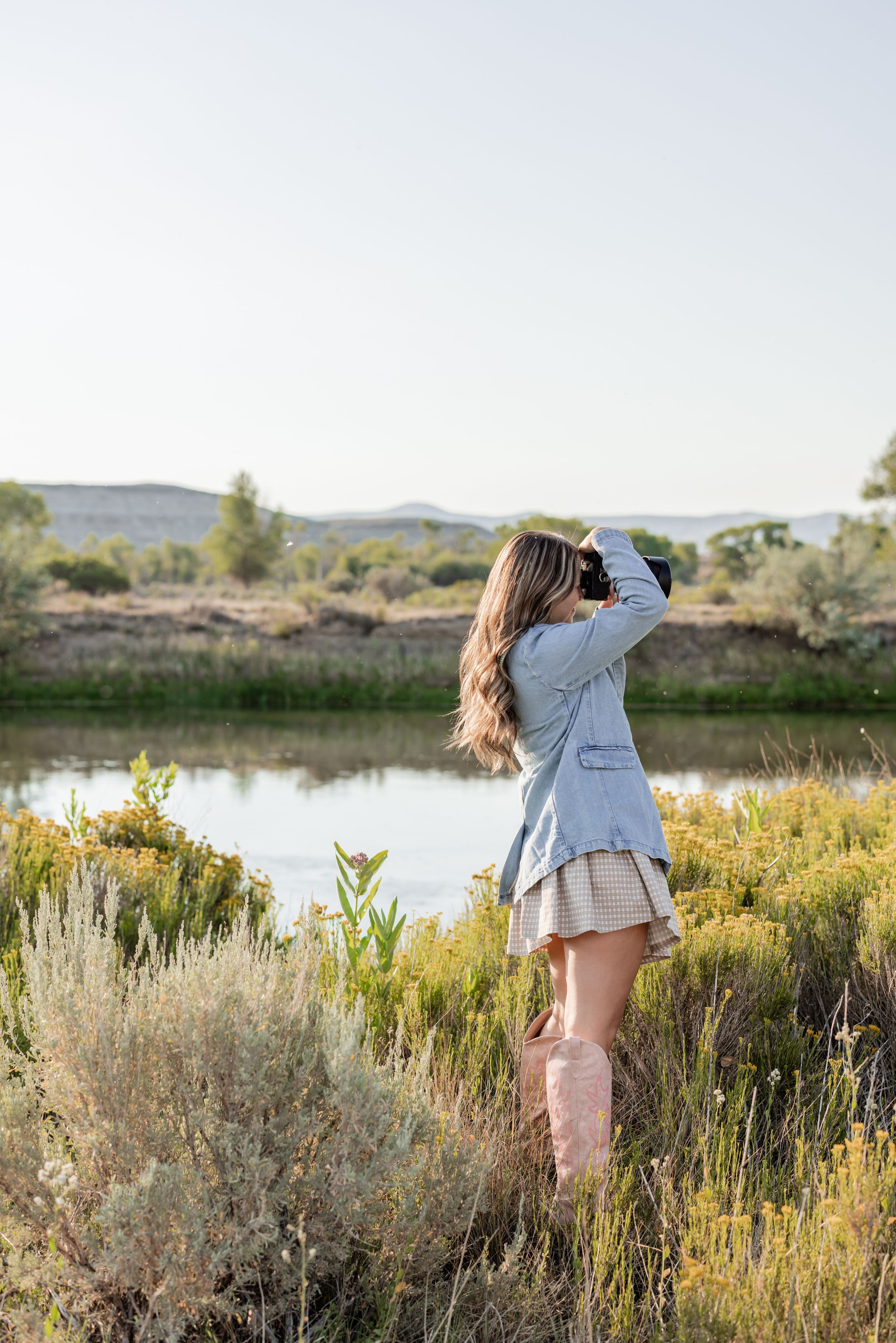 Woman in denim jacket, mini skirt, and pink boots photographing a serene river landscape surrounded by wildflowers.