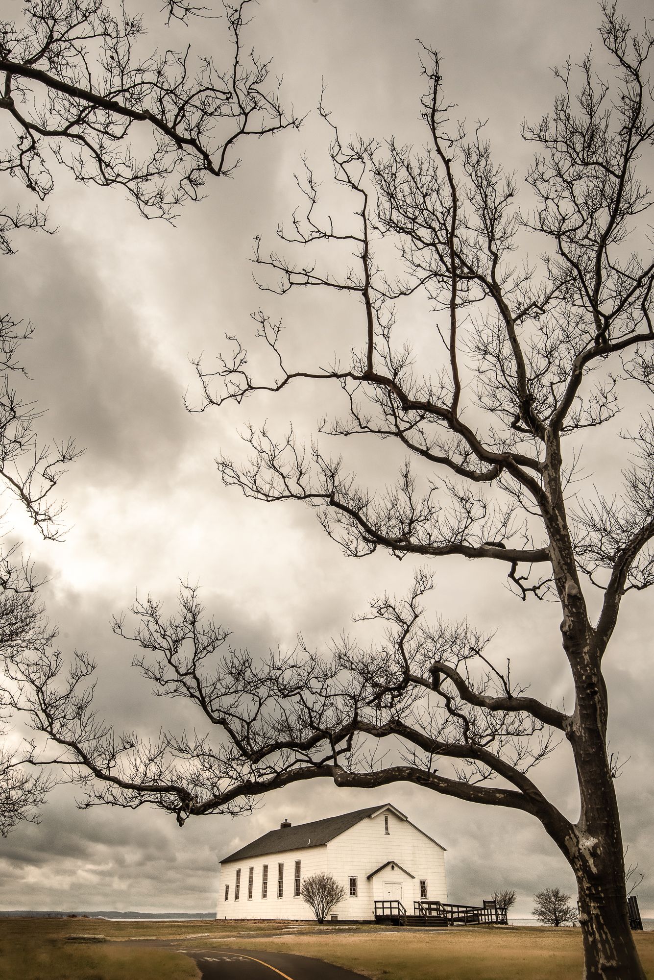 Dramatic black and white photo of a solitary church beneath twisted bare tree branches in Fort Hancock, Sandy Hook, NJ