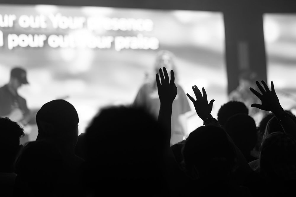 Silhouetted crowd at a concert with raised hands against a bright projection screen.