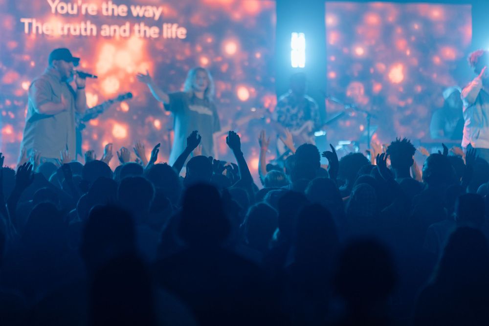 Concert crowd silhouetted against blue lighting with lyrics projected on screen.