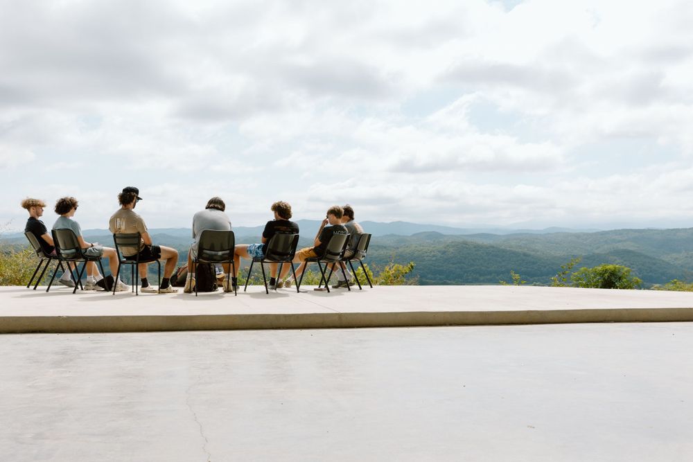 People sitting in chairs on a mountaintop platform overlooking a scenic valley view.