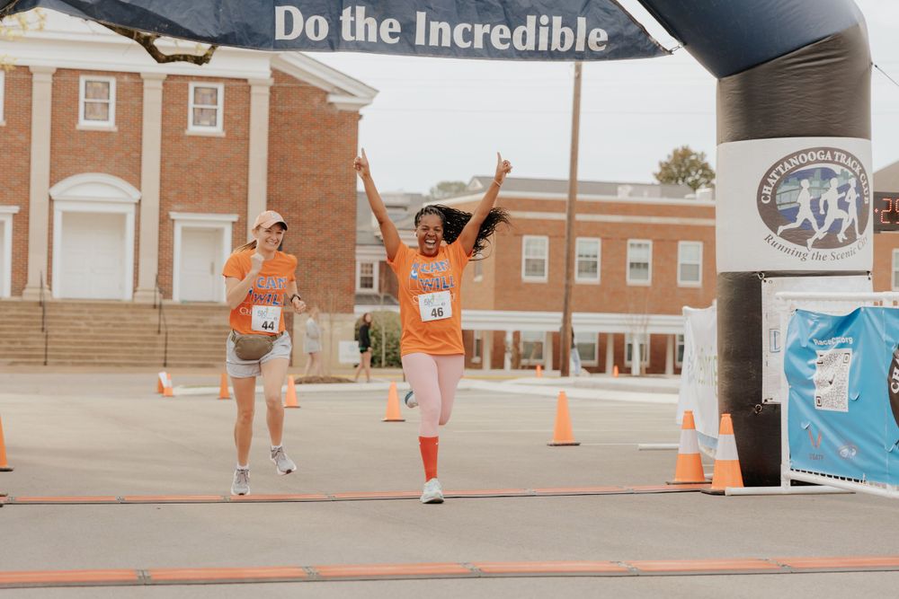 Two runners in bright orange shirts celebrate as they cross the finish line at a running event beneath an arch.