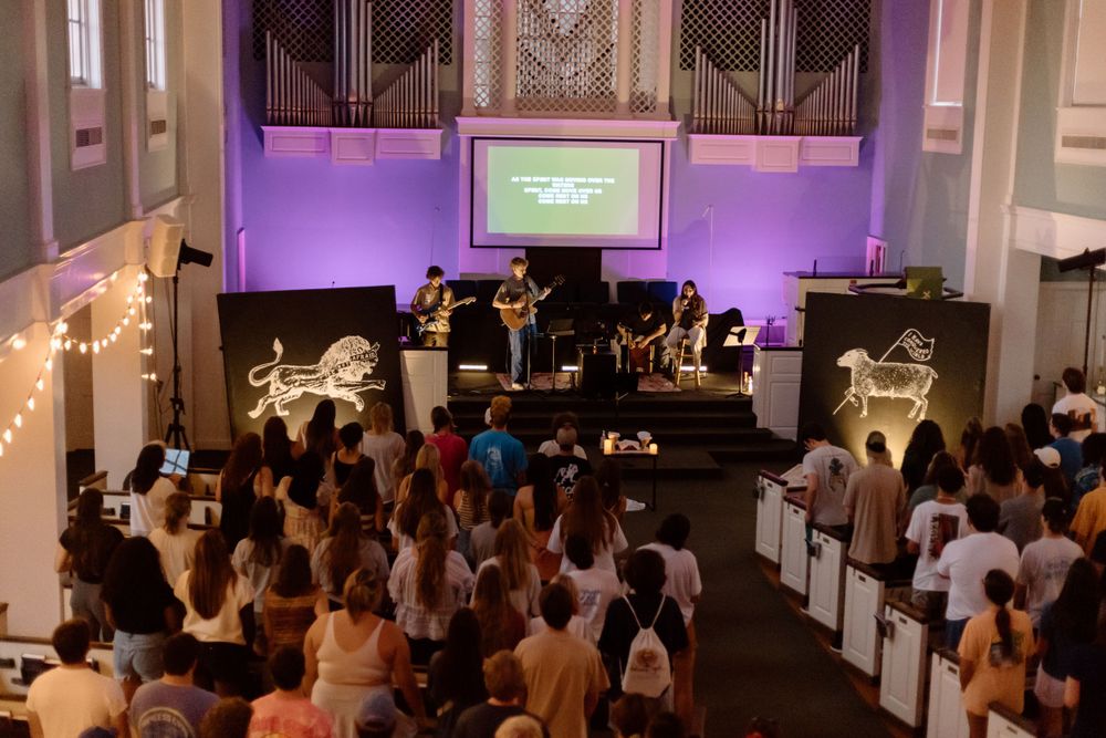 Church congregation gathers for worship service with purple stage lighting and projection screen.