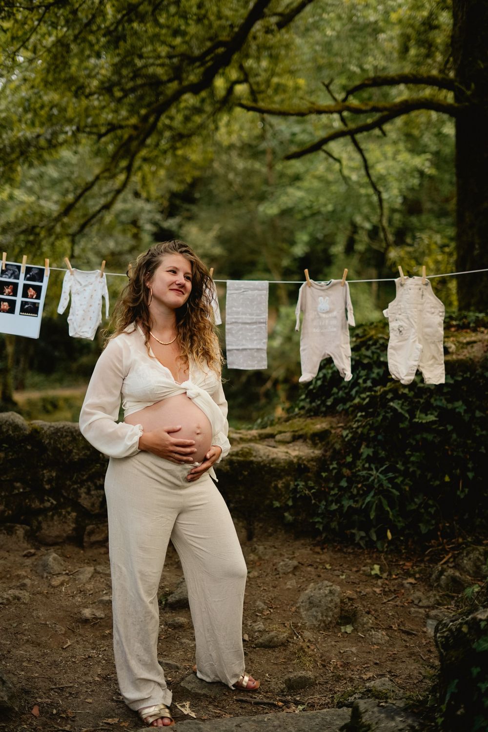 Pregnant woman in white outfit poses near baby clothes hanging on outdoor clothesline.