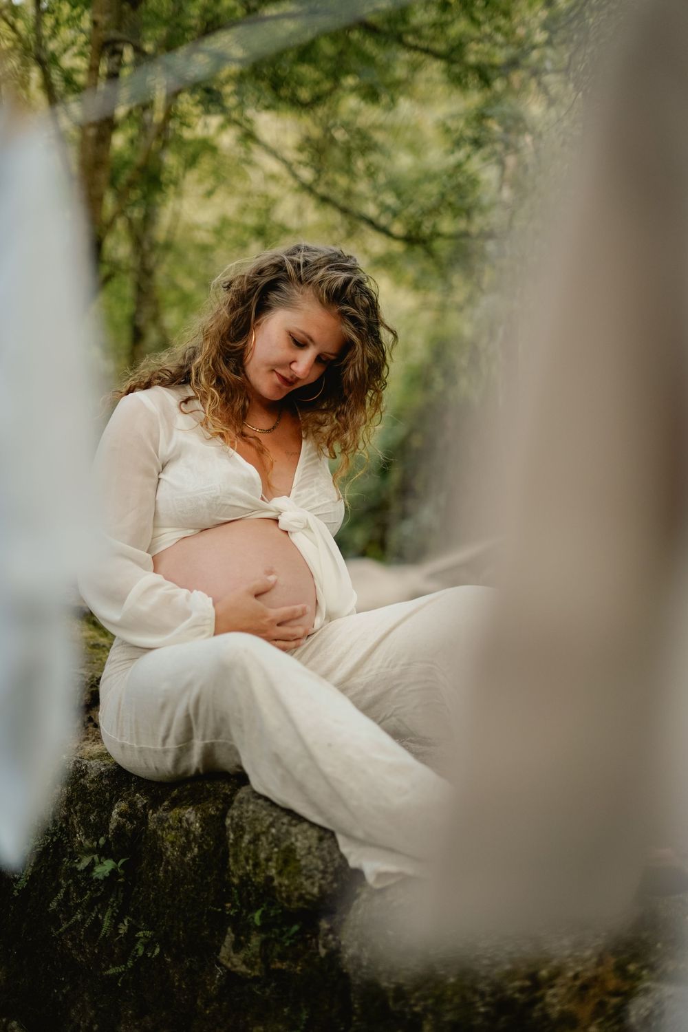 Maternity portrait of woman in white dress sitting on rock in forest setting.
