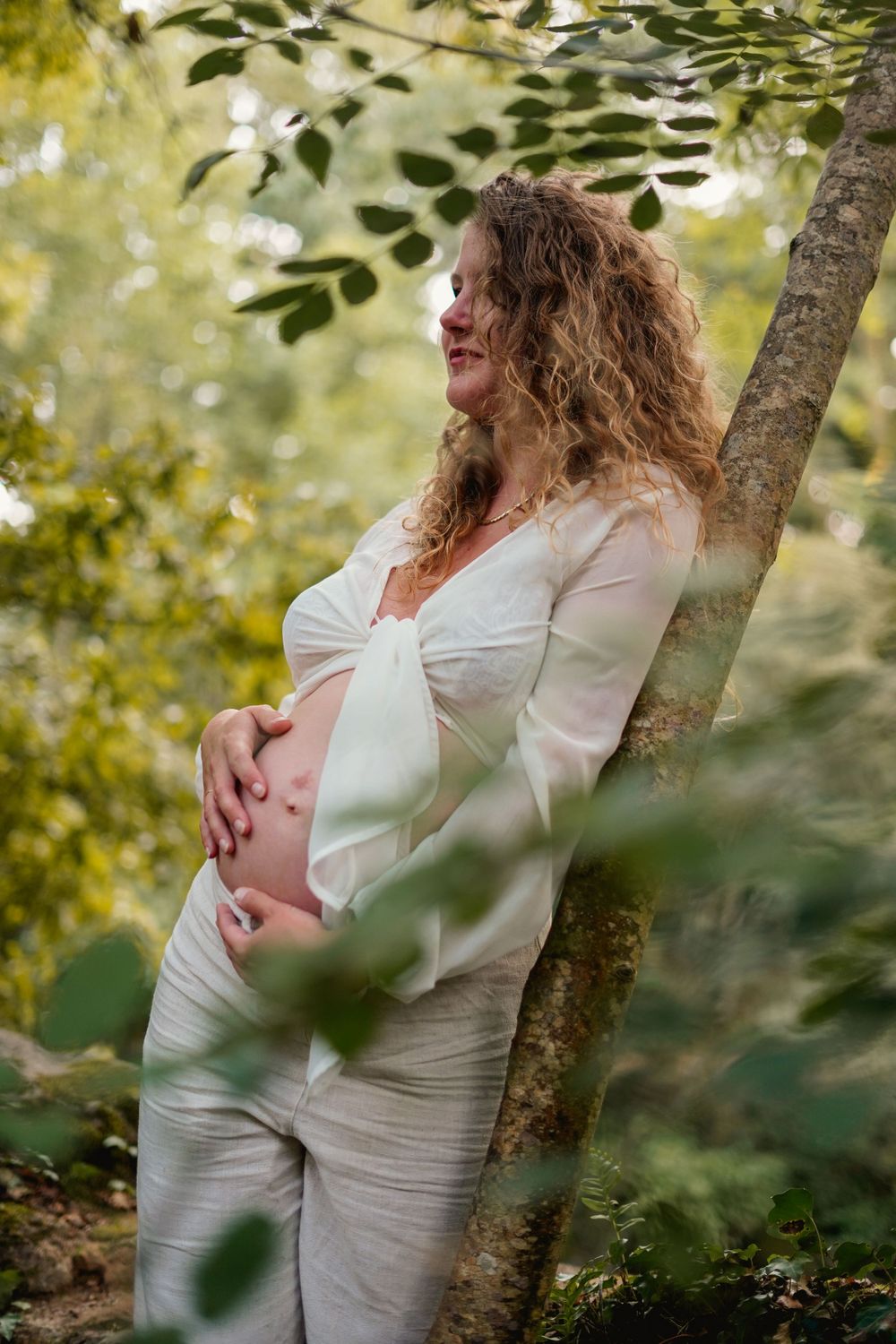 Artistic maternity photo in nature showing an expectant mother in white leaning against a tree in soft sunlight.