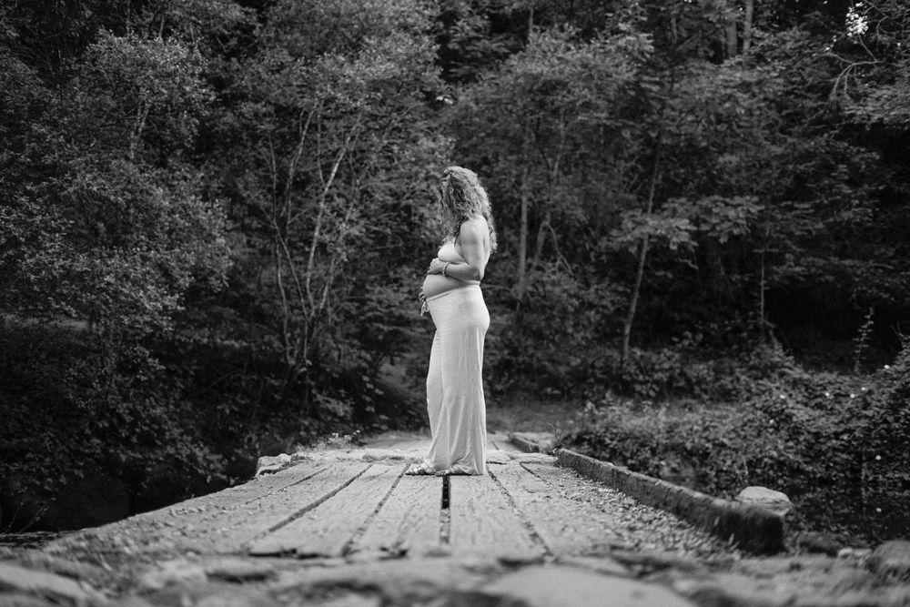 Black and white maternity silhouette standing on wooden bridge in forest setting.