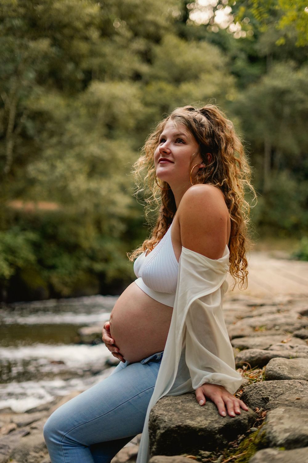 Outdoor maternity portrait by river rocks showing pregnant figure in white top and jeans.