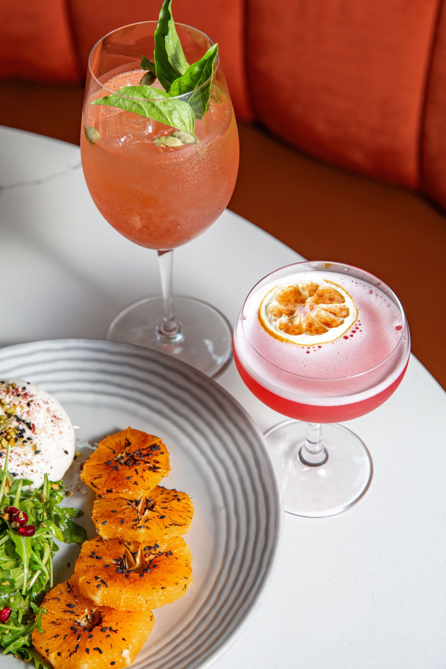 Orange-colored cocktails garnished with citrus and herbs served on patterned plates.