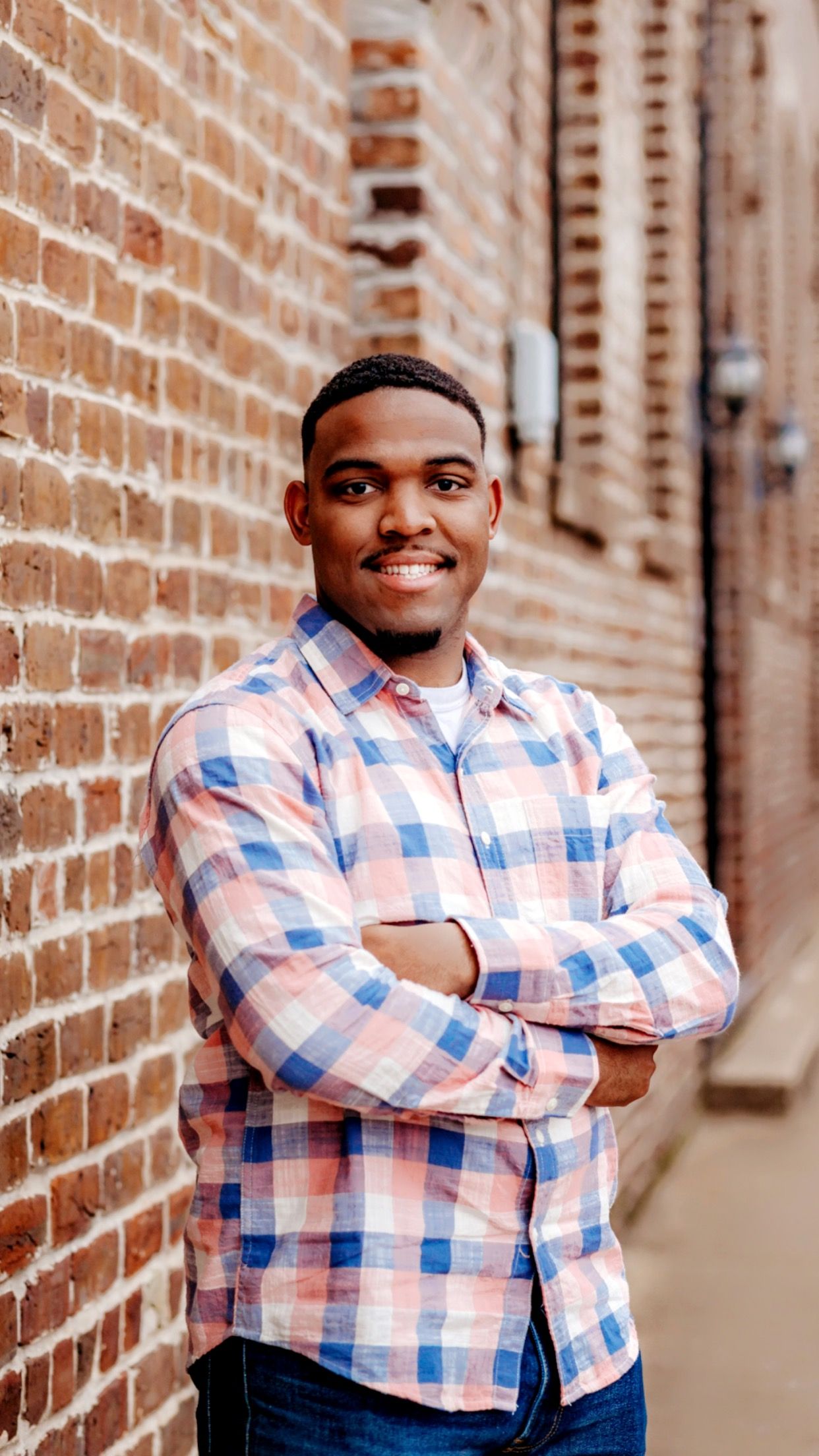 Someone wearing a pink and blue plaid button-down shirt poses confidently against a brick wall in an urban setting.
