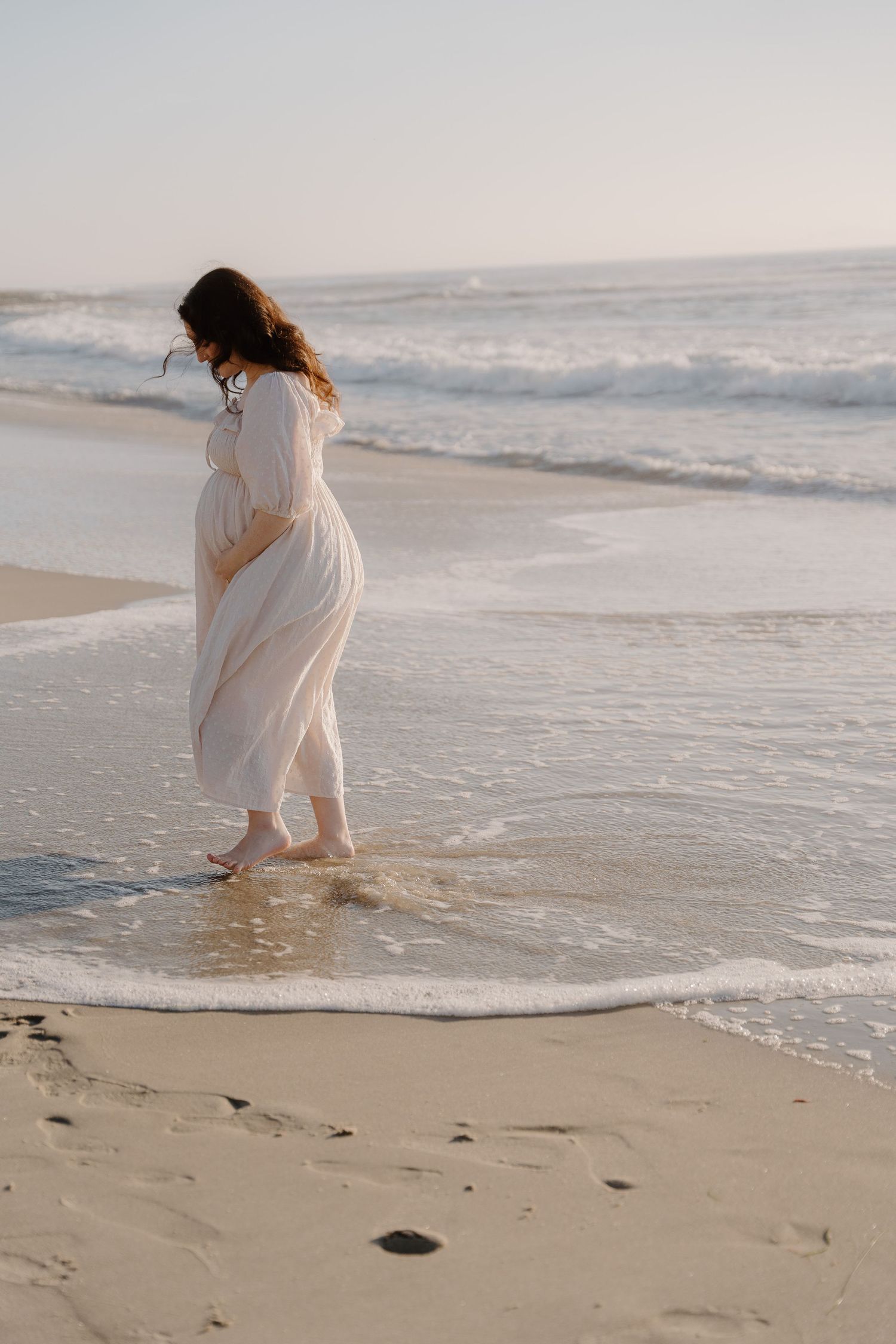 A pregnant woman with a flowing white dress that catches the breeze as waves roll onto a sunset beach at La Jolla beach in San Diego, CA.