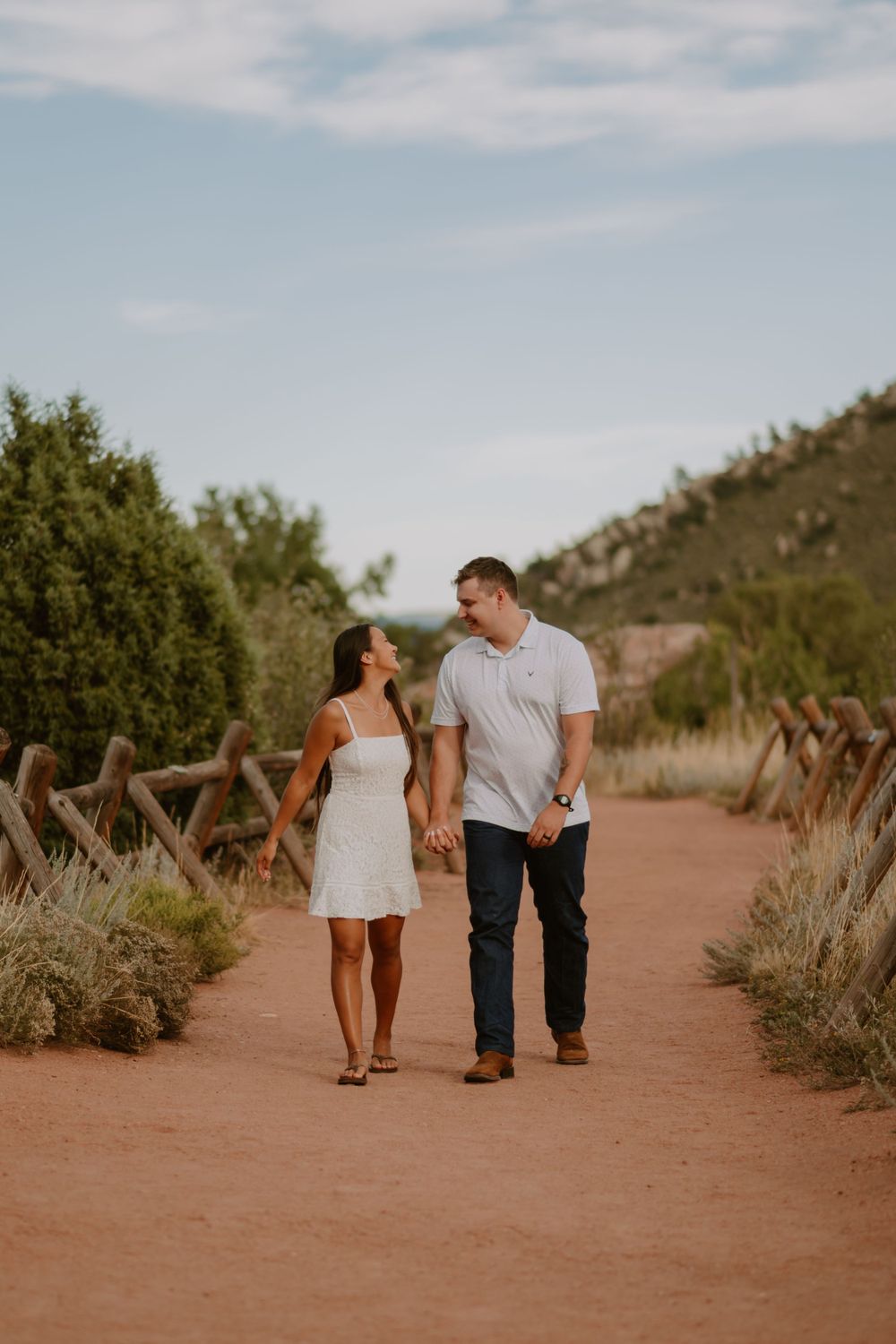 A couple holding hands and smiling at each other while walking on a dirt path surrounded by greenery and hills.