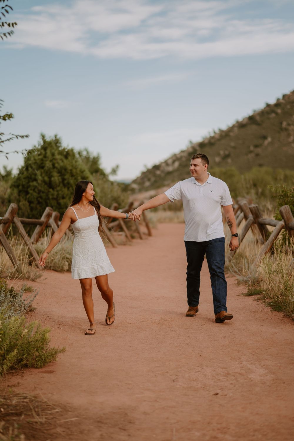 A couple holding hands, walking on a dirt path surrounded by greenery and mountains under a partly cloudy sky.