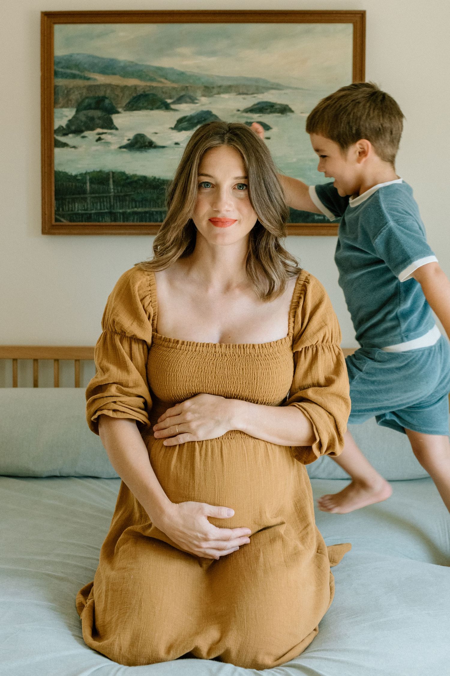 Expecting mother in mustard dress sitting on bed with coastal artwork in background during family maternity photoshoot in San Diego.